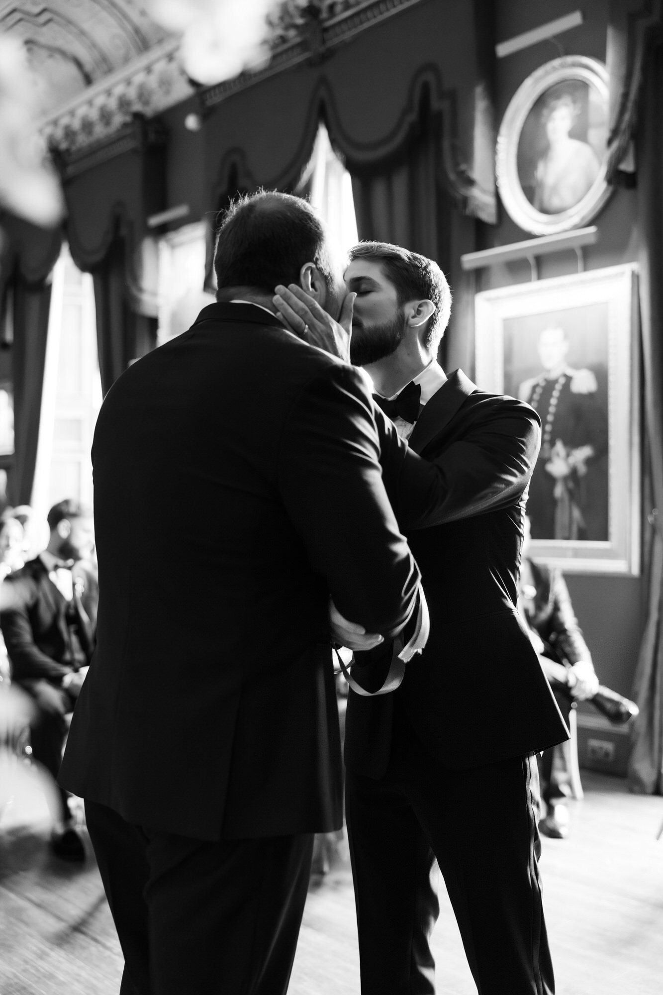 A black and white photograph of two men in tuxedos sharing a kiss during a formal event, possibly a wedding, inside an ornately decorated room with framed portraits on the wall.
