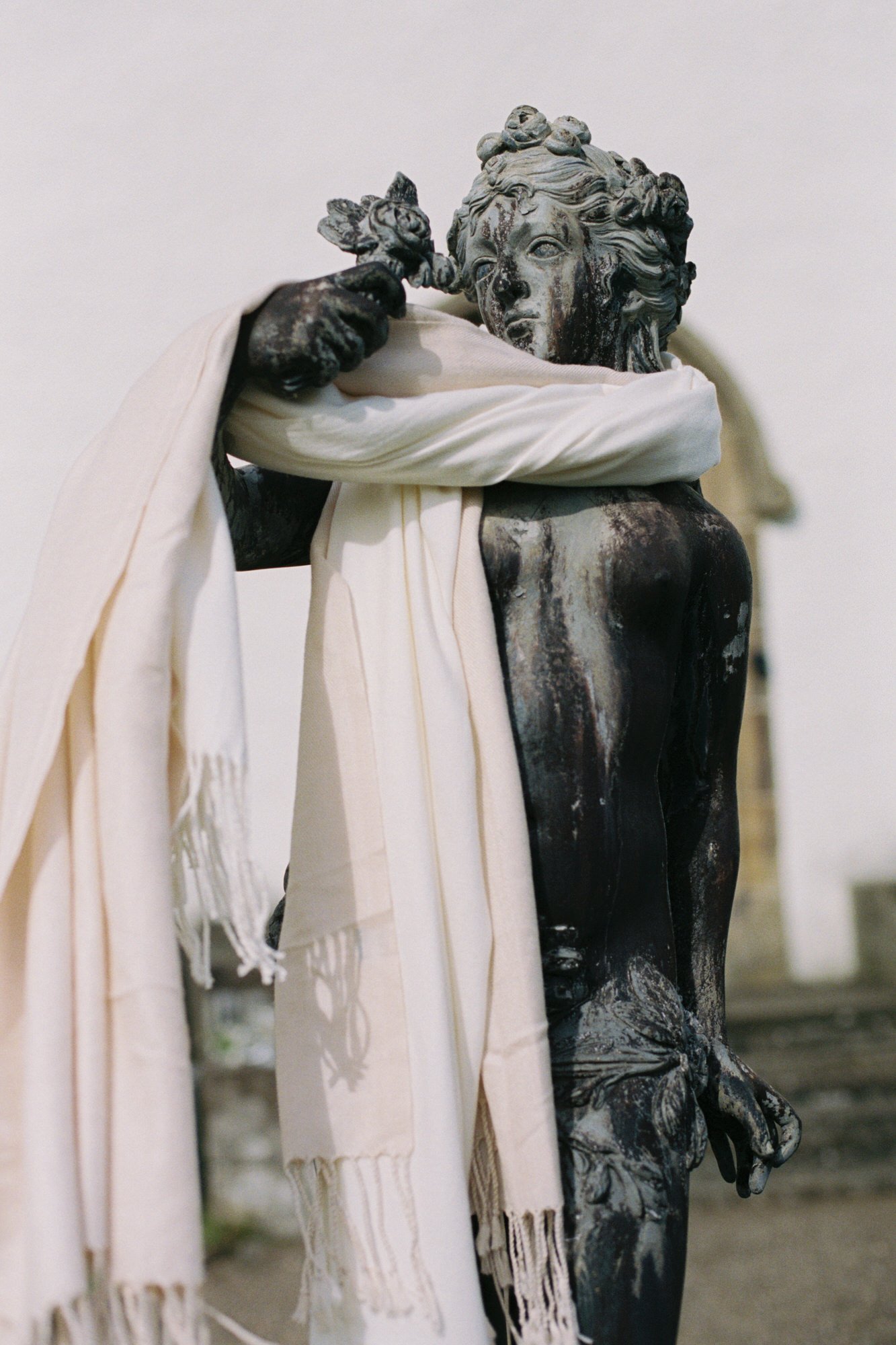 A weathered, blackened classical sculpture of a male figure with curly hair, holding a cloth to his shoulder, with white fabric draped over his left arm. Photographed by Steven William Weir, Edinburgh Scotland.