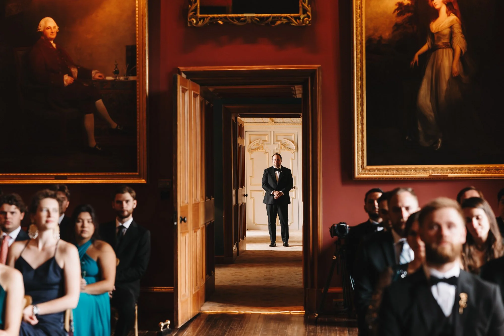 A formal event in an elegant room with red walls and large framed portraits. People are seated, dressed in formal evening wear. A man in a tuxedo stands in the doorway, looking into the room.