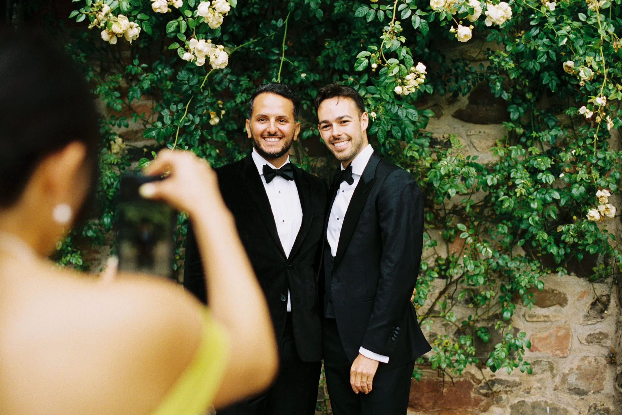 Two men in tuxedos posing for a photo in front of a stone wall covered with green plants and white flowers, as a woman in a yellow dress takes their picture. Photographed by Steven William Weir, Edinburgh Scotland.
