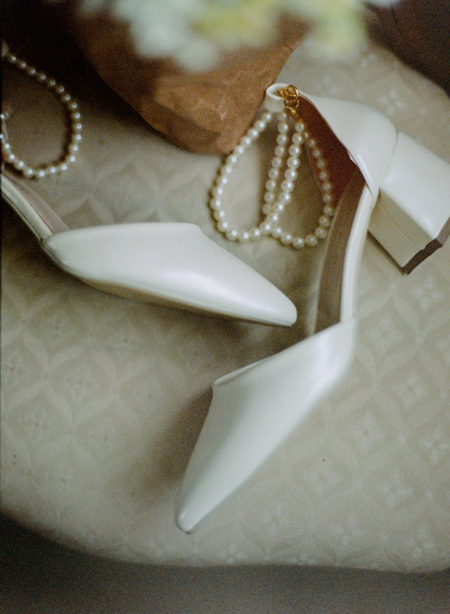 A pair of white high-heeled shoes, pearl necklaces, and a brown paper bag on a patterned surface. Photographed by Steven William Weir, Edinburgh Scotland.