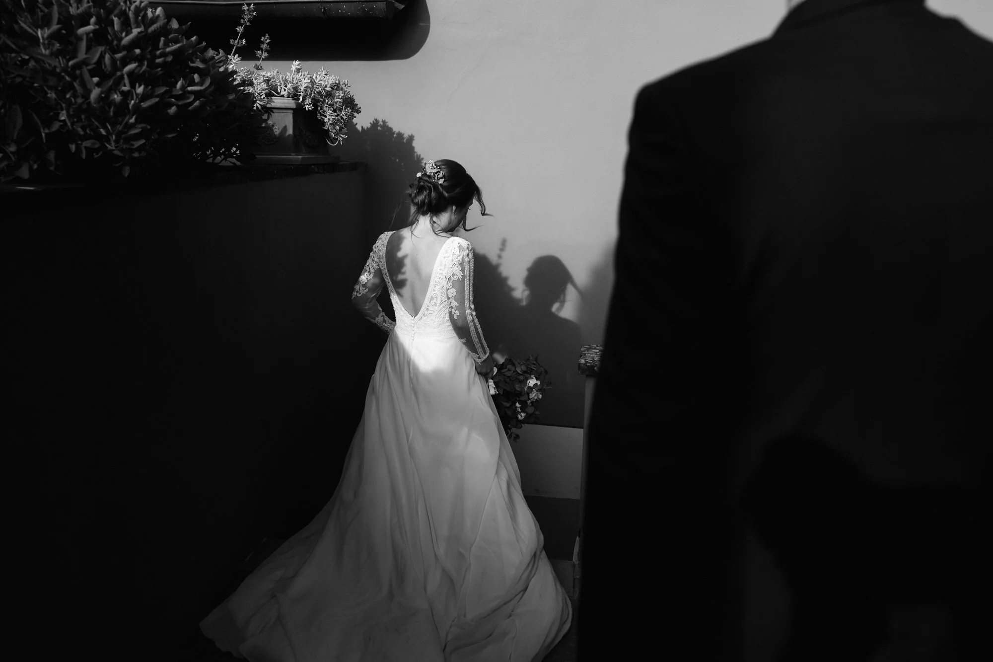 A black and white photo of a bride in a wedding dress with lace sleeves, holding a bouquet, standing with her back to the camera. The bride has her hair in an updo and is illuminated by a light source creating a shadow on the wall. A man in a suit is