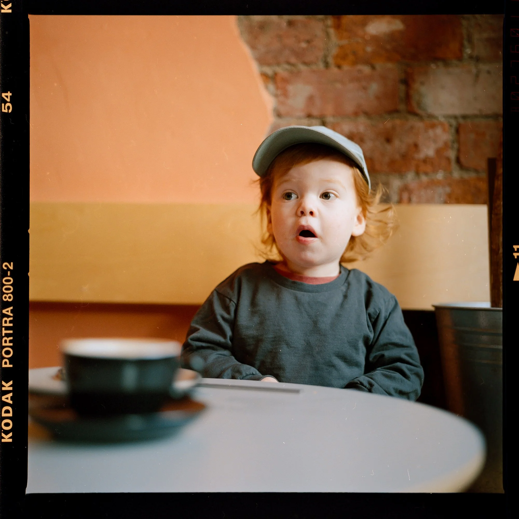 A young boy with red hair, wearing a gray cap and a dark gray shirt, sitting at a table with surprised or curious expression, with a blurred cup in the foreground and a brick wall in the background.