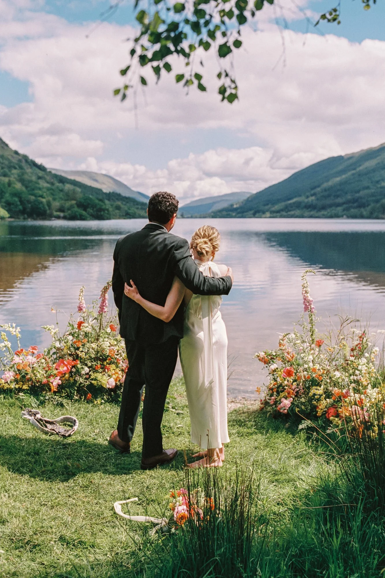 A couple in wedding attire embracing by a lakeside, with mountains and a cloudy sky in the background, surrounded by colorful flowers. Photographed by Steven William Weir, Edinburgh Scotland.