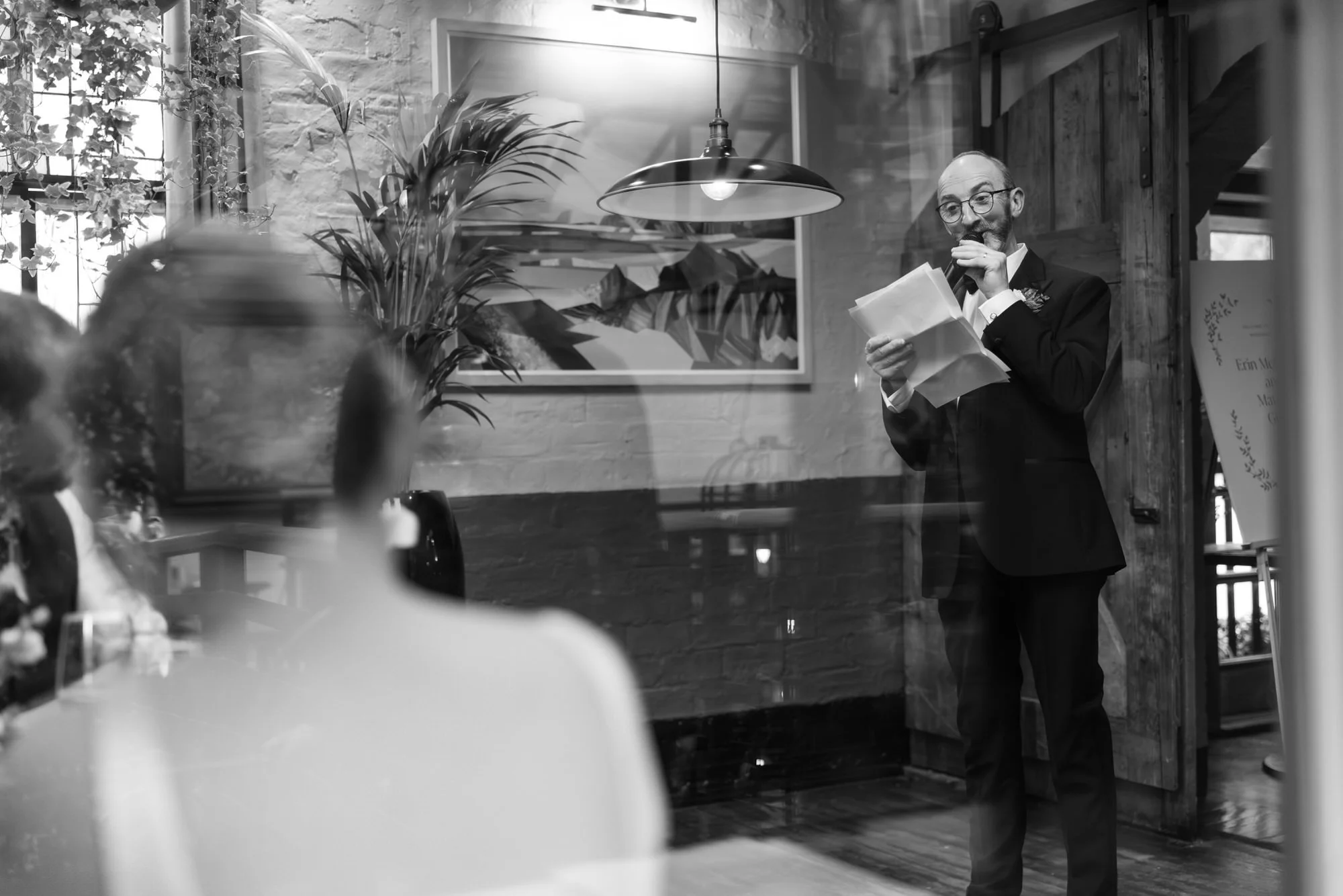 A man in a suit giving a speech or toast at a wedding reception, with guests seated and listening. The setting is a cozy, decorated indoor space with artwork and a large plant. Photographed by Steven William Weir, Edinburgh Scotland.