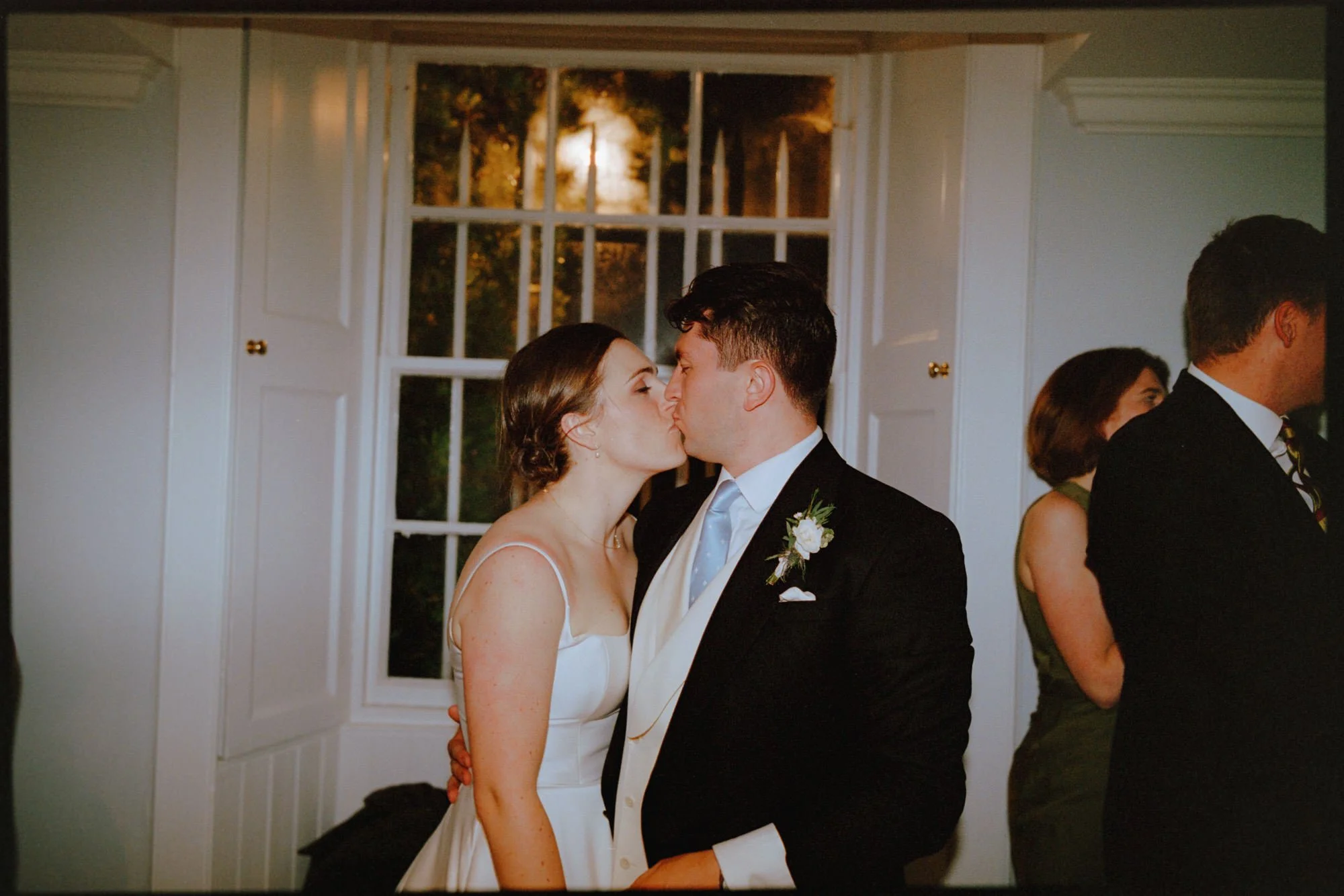 A bride and groom sharing a kiss at their wedding reception, with two guests in the background and a window with closed shutters behind them.