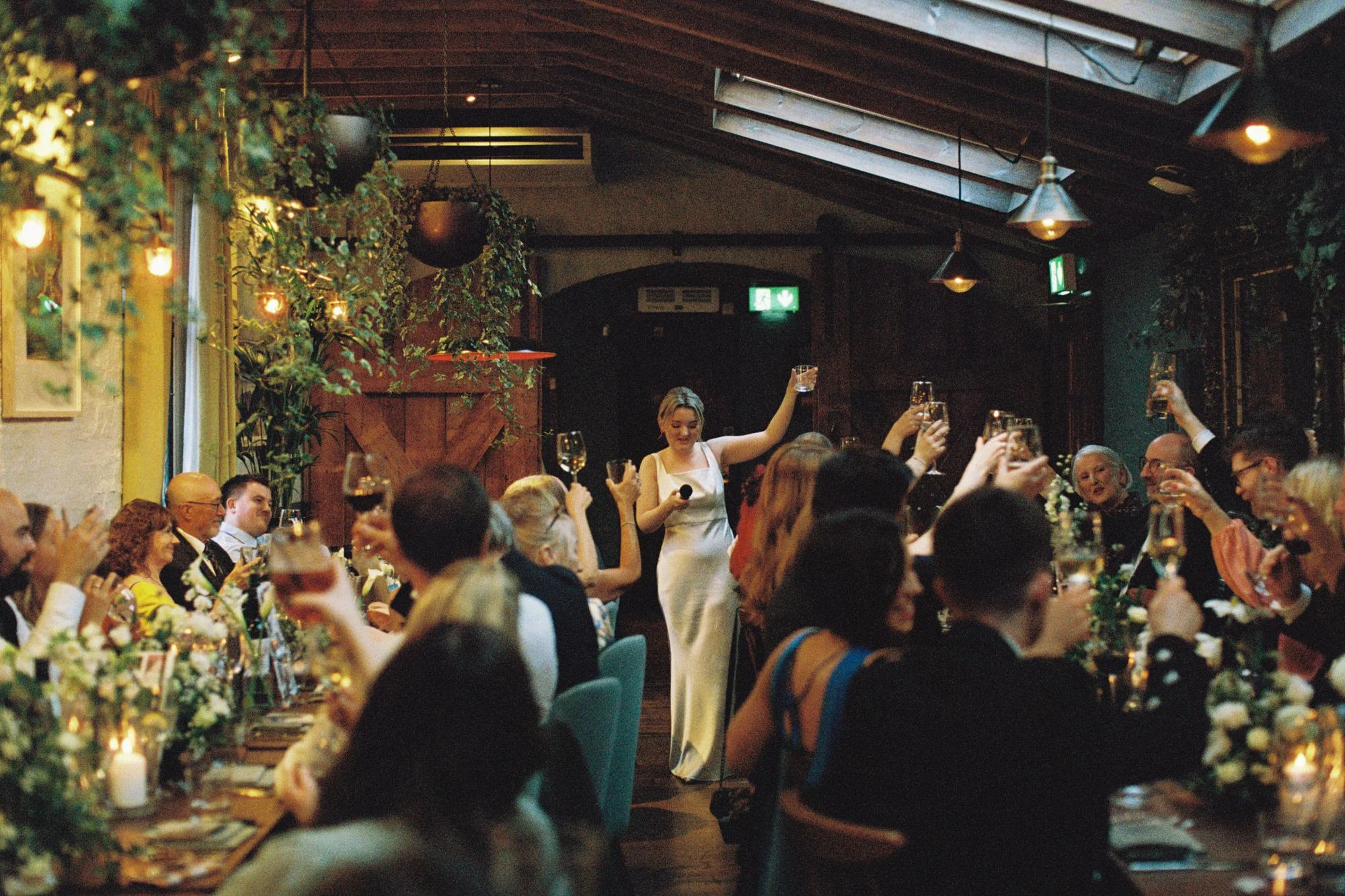A woman in a white dress giving a toast at a dinner party with guests at long tables, raising glasses in celebration, in a warmly lit, decorated restaurant setting. Photographed by Steven William Weir, Edinburgh Scotland.