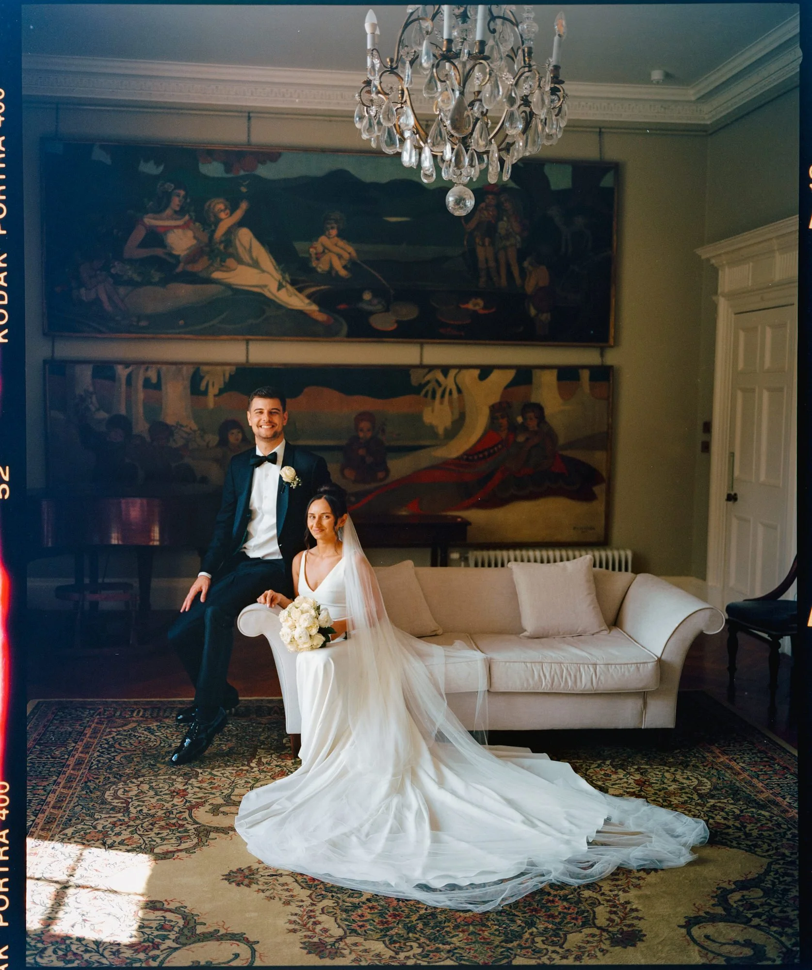 A bride and groom in wedding attire sitting on a white couch in a decorated room with paintings on the wall, a chandelier hanging from the ceiling, and a patterned rug on the floor. Photographed by Steven William Weir, Edinburgh Scotland.