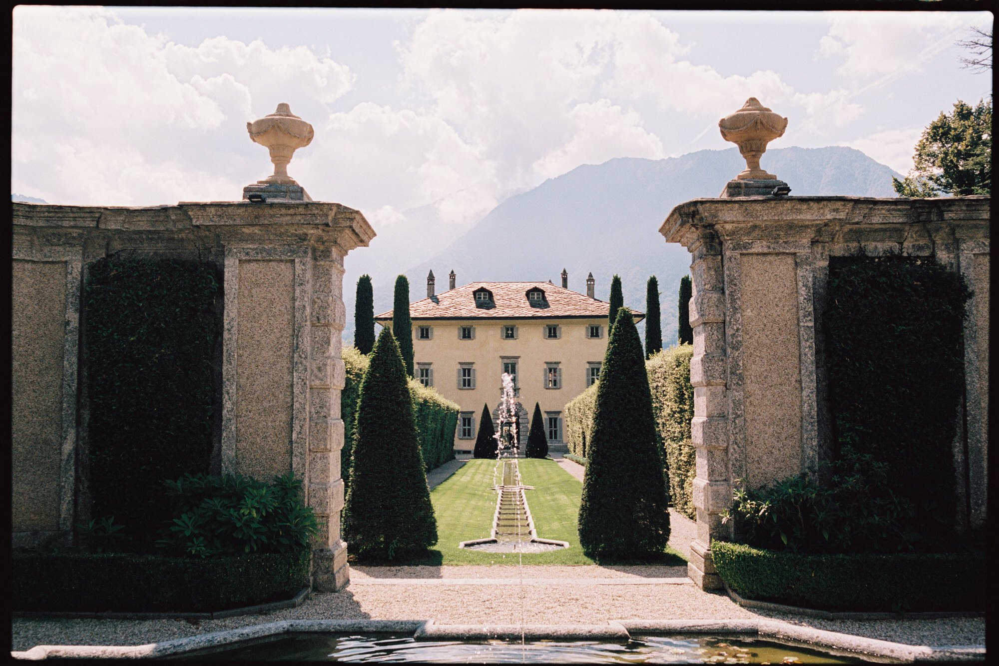 View of a grand estate, Villa Balbiano with a formal garden, fountain, and a large house in the background, framed by stone pillars and lush greenery, with mountains in the distance and partly cloudy sky.