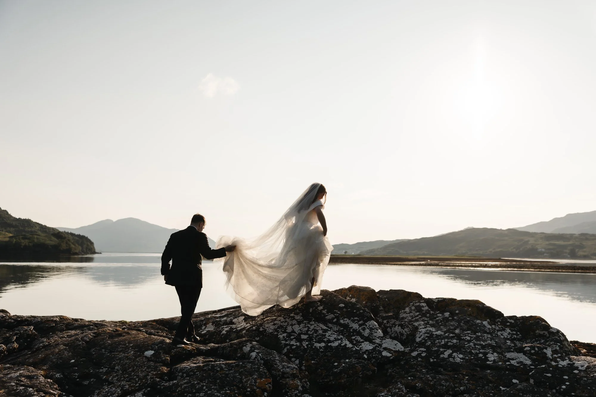 Bride in a white wedding dress and veil walking on rocks near a lake with a groom in a black suit holding her dress train at sunset.