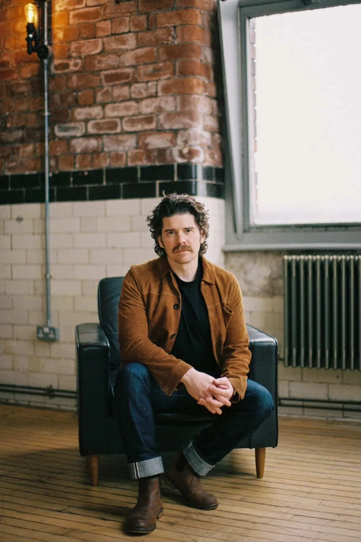A man with dark, curly hair and a mustache sitting cross-legged on a black armchair in a room with exposed brick wall and a large window.