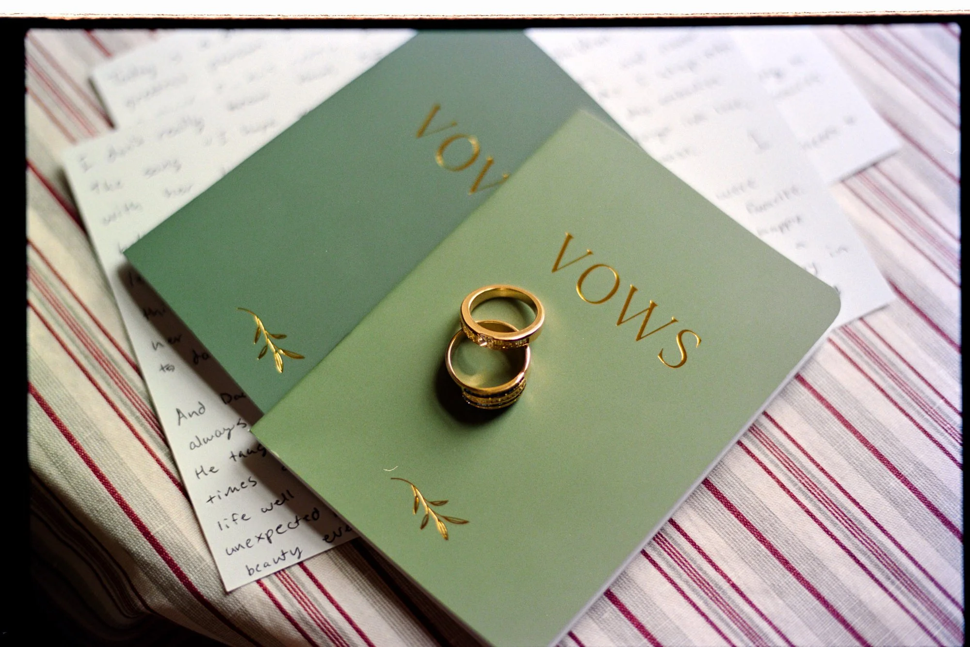 Green wedding program with gold lettering that says 'Vows' and gold decorative accents, with three wedding rings placed on top, sitting on a striped tablecloth with handwritten notes in the background.