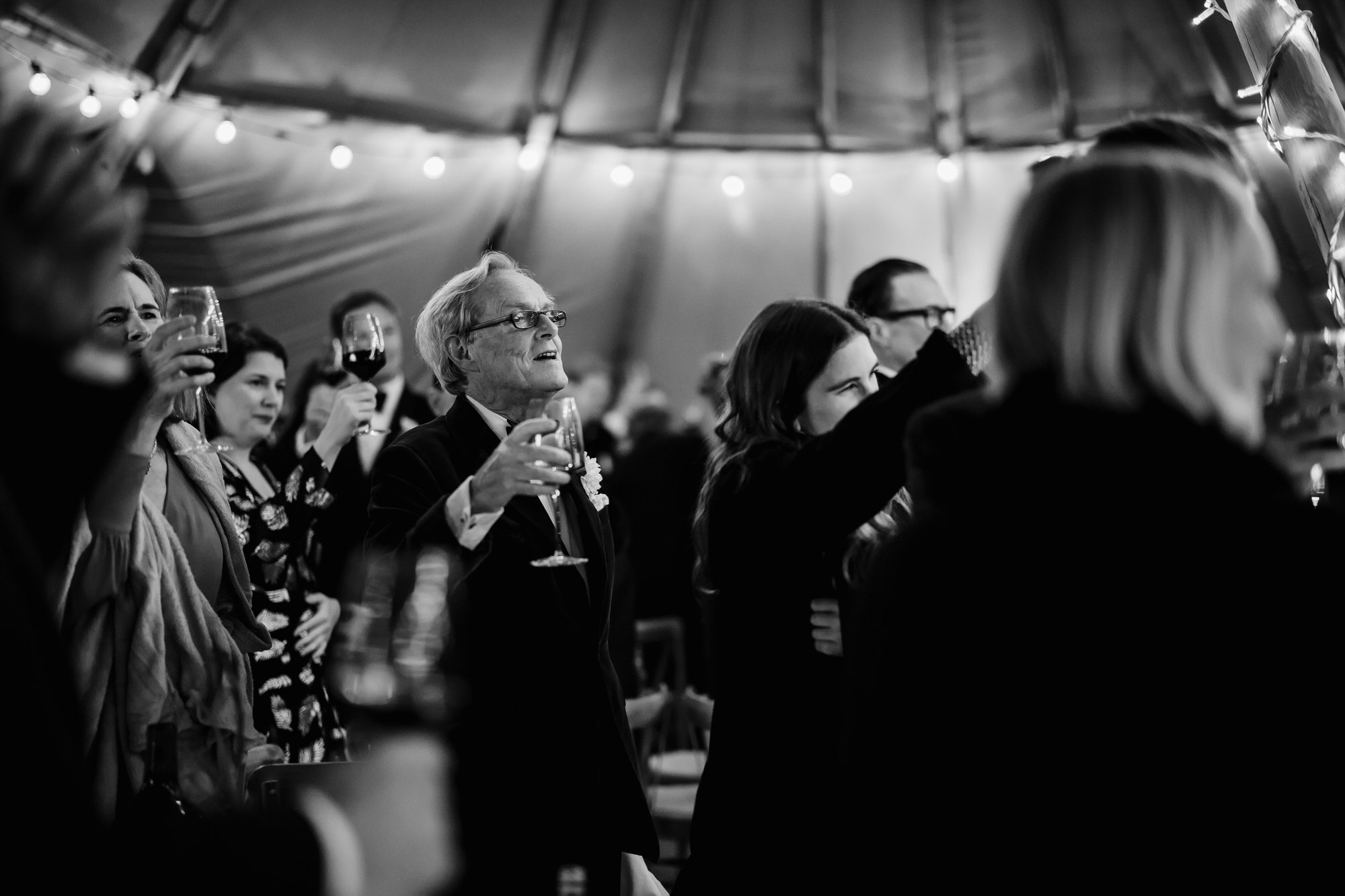 Black and white photo of people at a celebration raising wine glasses, some smiling and looking up, under a tent with string lights. Photographed by Steven William Weir, Edinburgh Scotland.