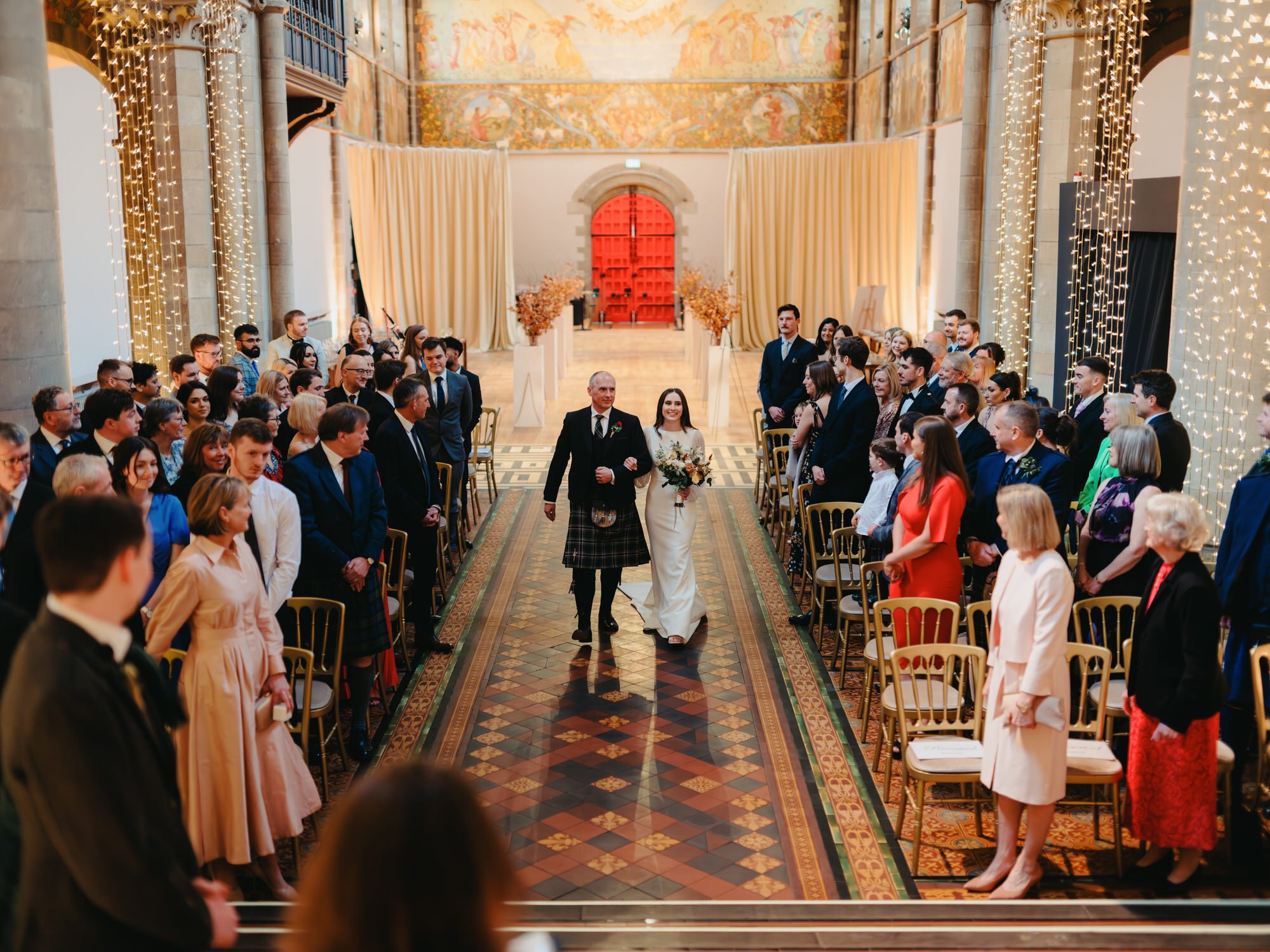 Bride walking down the aisle with her father in a decorated indoor wedding venue, surrounded by seated guests. Photographed by Steven William Weir, Edinburgh Scotland.