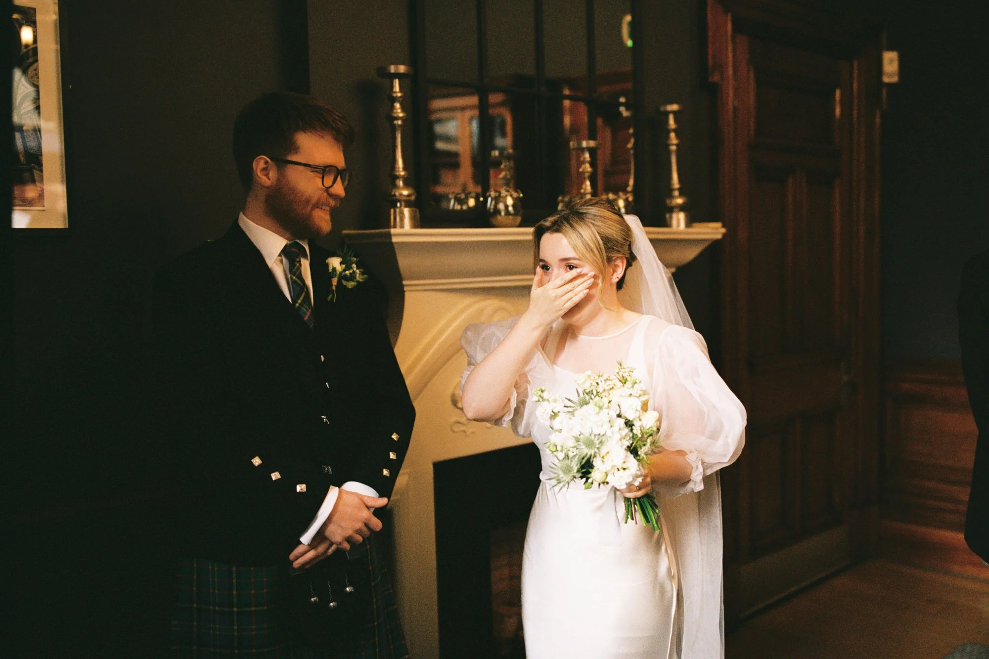 A bride in a white wedding dress holding a bouquet of white flowers, covering her mouth with her hand and looking surprised, stands next to a groom in a dark suit with a tartan kilt, who is smiling during a wedding ceremony indoors with dark wood pan
