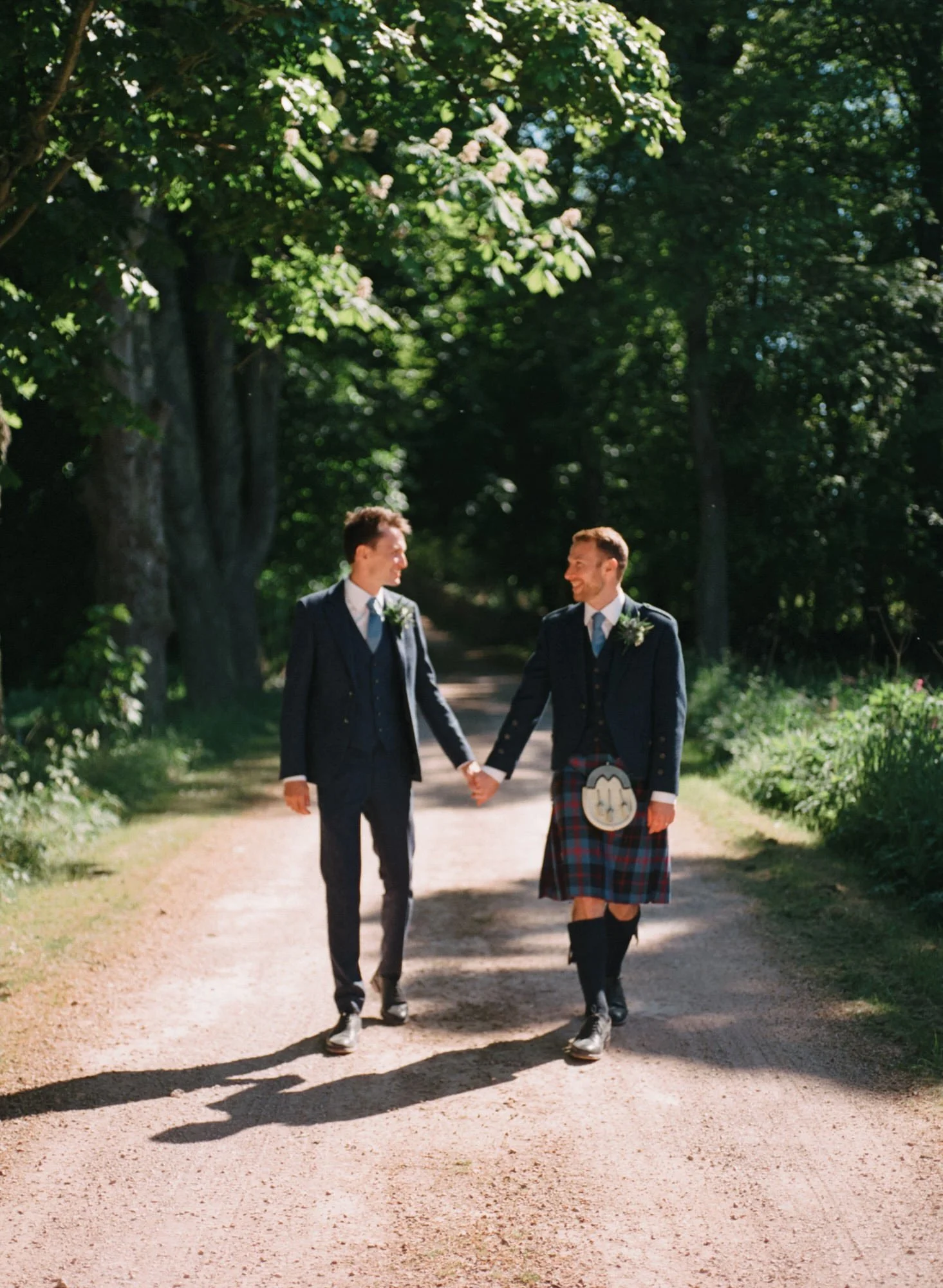 Two men holding hands and walking on a sunlit forest path, dressed in formal attire; one wears a kilt with a sporran.