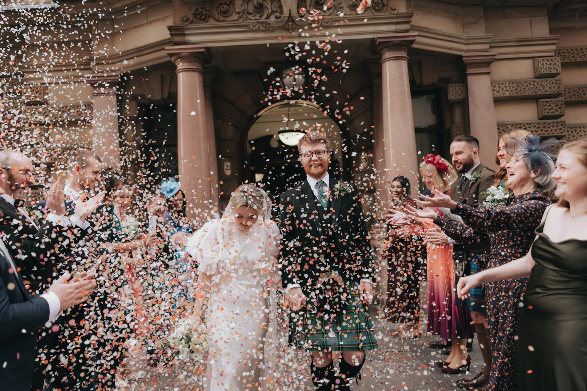 A newly married couple walking through a shower of colorful confetti, surrounded by friends and family outside a grand building with stone columns.