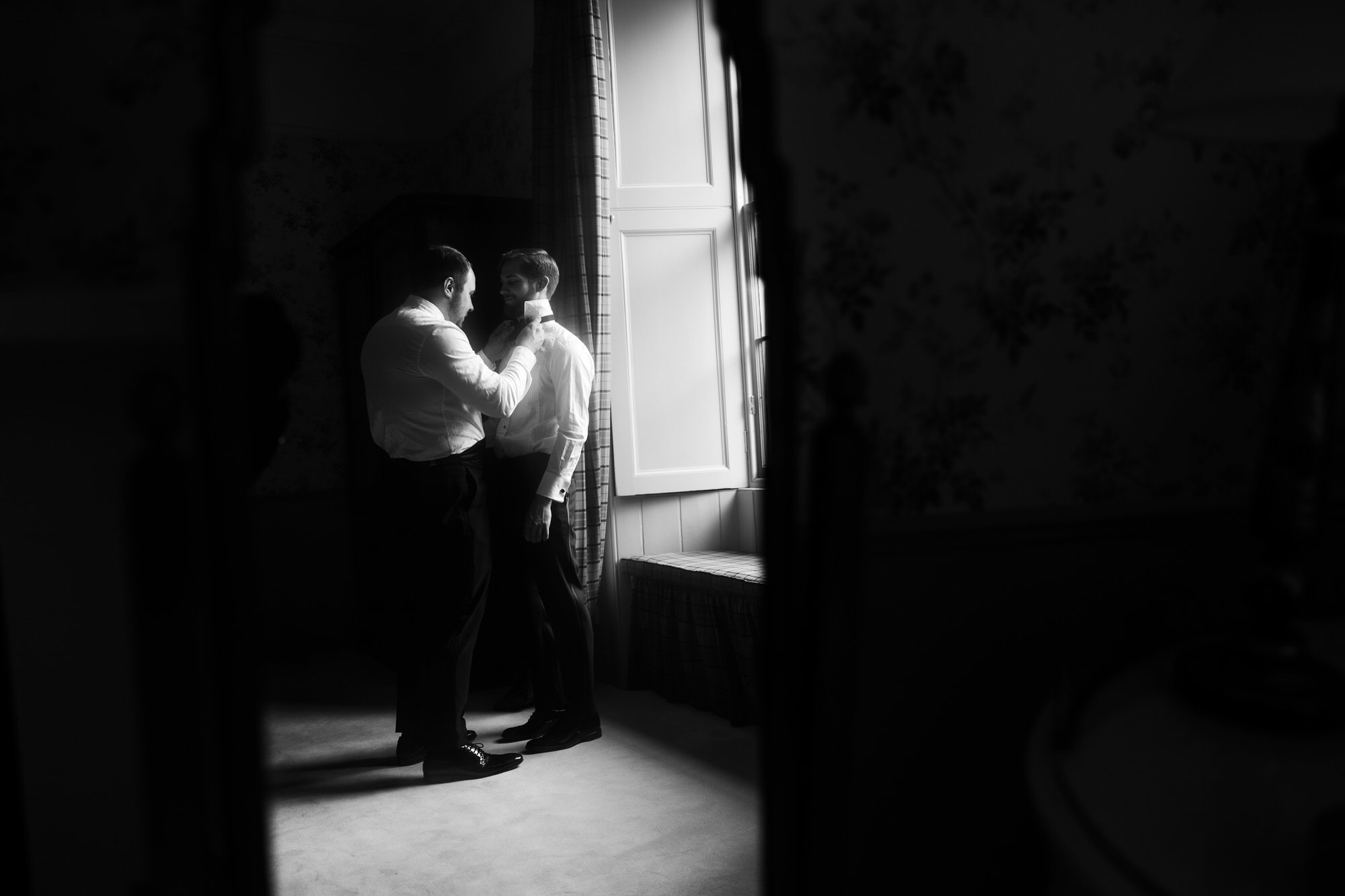 Two men in formal dress inside a room, one adjusting the other's collar near a window.