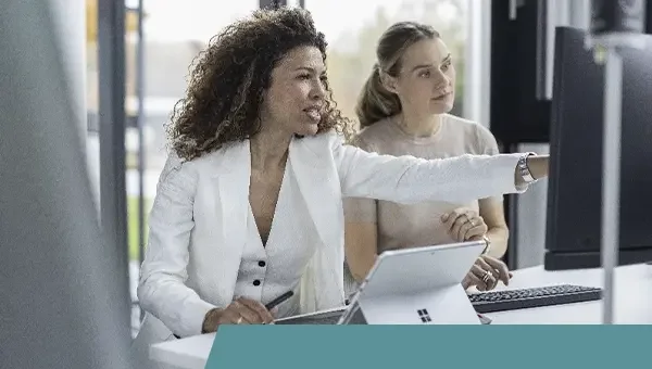 Zwei Frauen in einem Büro arbeiten an Computern, eine erklärt etwas und zeigt auf den Bildschirm.