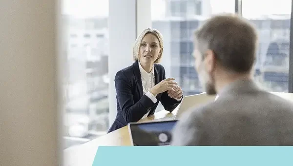 Zwei Menschen in einem Büro bei einem Gespräch, eine Frau spricht, ein Mann hört zu, im Hintergrund Fenster mit Stadtansicht.