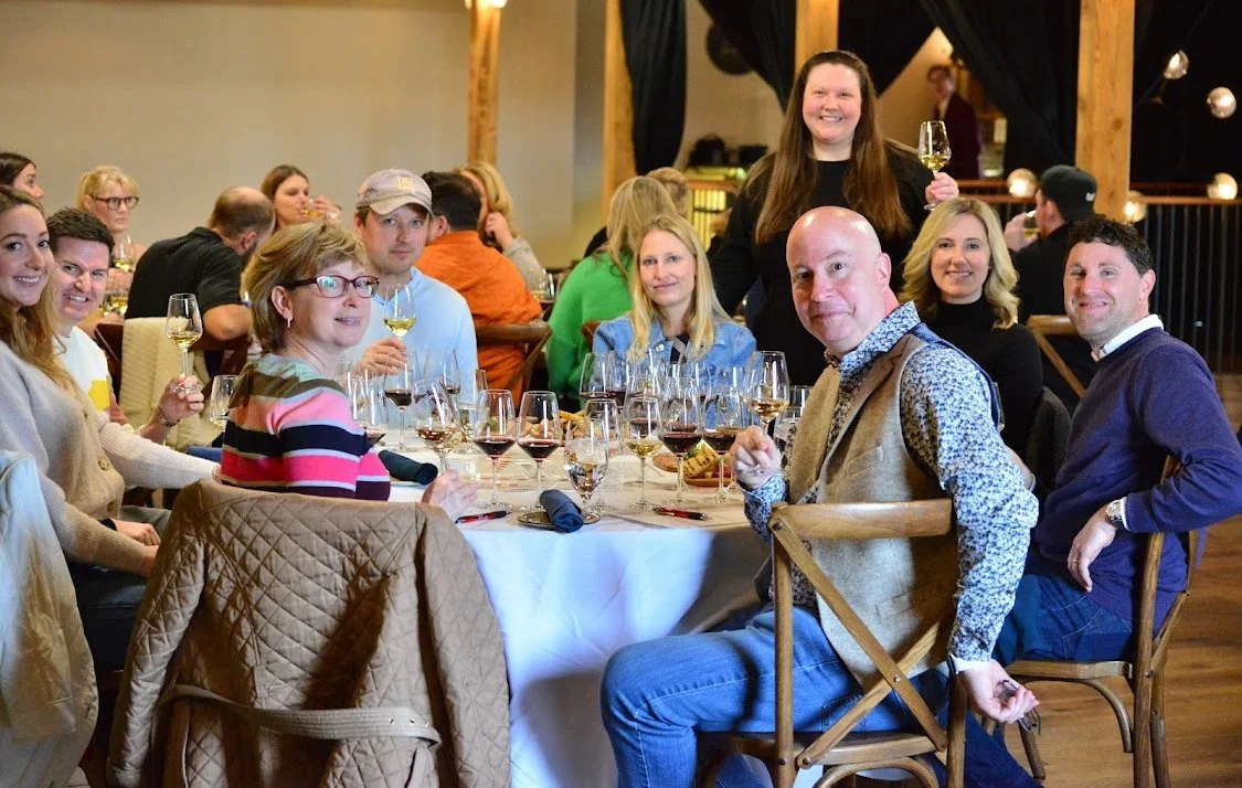 scene from a private wine tasting. A group of guests sits at a long table with flights of wine in front of them, while Amy stands behind the table, smiling for the camera.