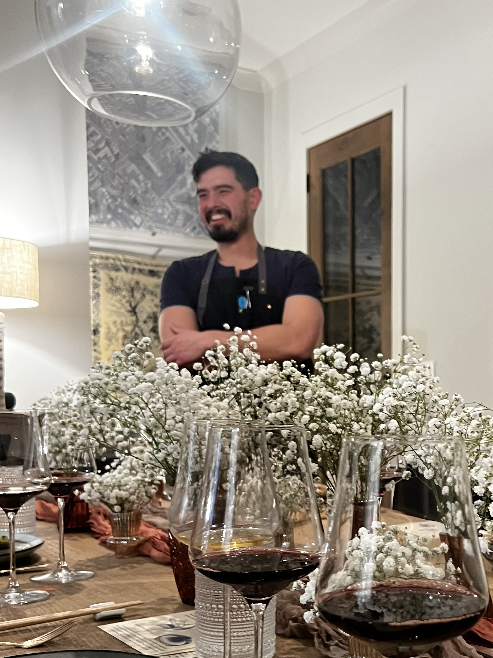 Chef Michael I stands at an elegant private dining table, smiling warmly. The table is decorated with white delicate florals and a sea of wine glasses.