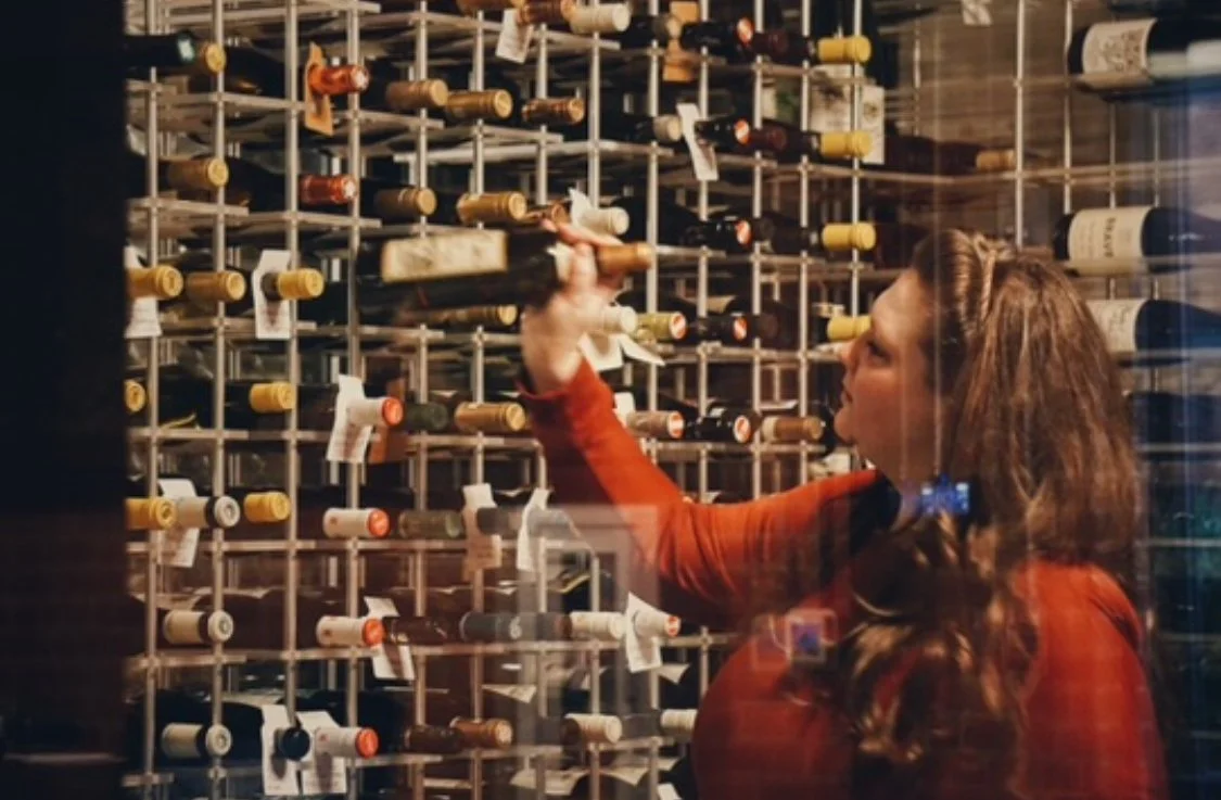 An atmospheric, artistic photo of Sommelier Amy in a dimly lit wine cellar. She is carefully selecting a bottle from a row of wines resting on their sides