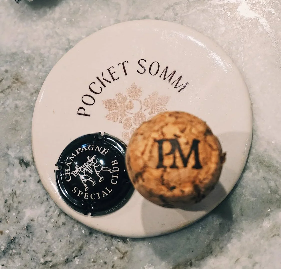 A close-up, top-down view of a Pocket Somm branded coaster. Resting on it is a Champagne a  cork and muselet cap.