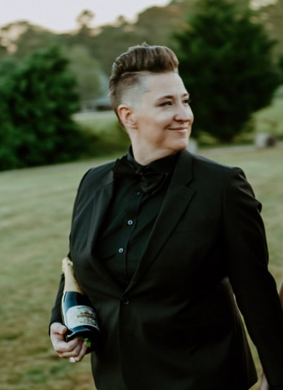 A professional portrait of Sommelier Christy K. She is posing confidently, resting a bottle of premium Champagne in the crook of her arm.