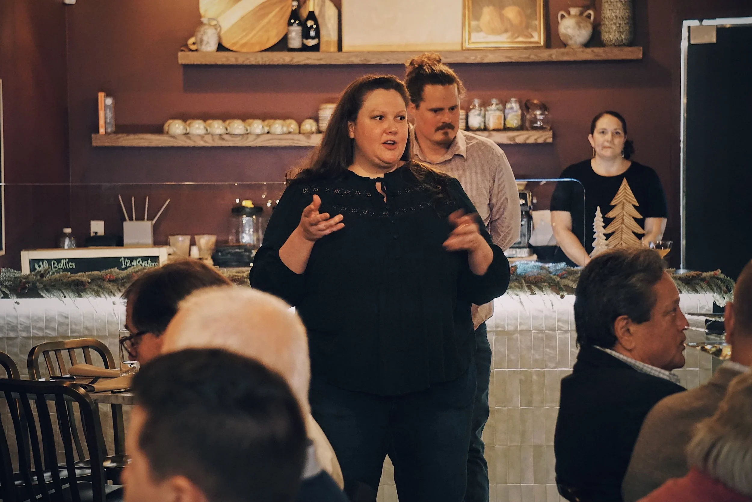 Sommelier Amy leading an elegant wine dinner. She stands confidently in a sophisticated dining room, sharing the story of a wine pairing with a room full of attentive guests.