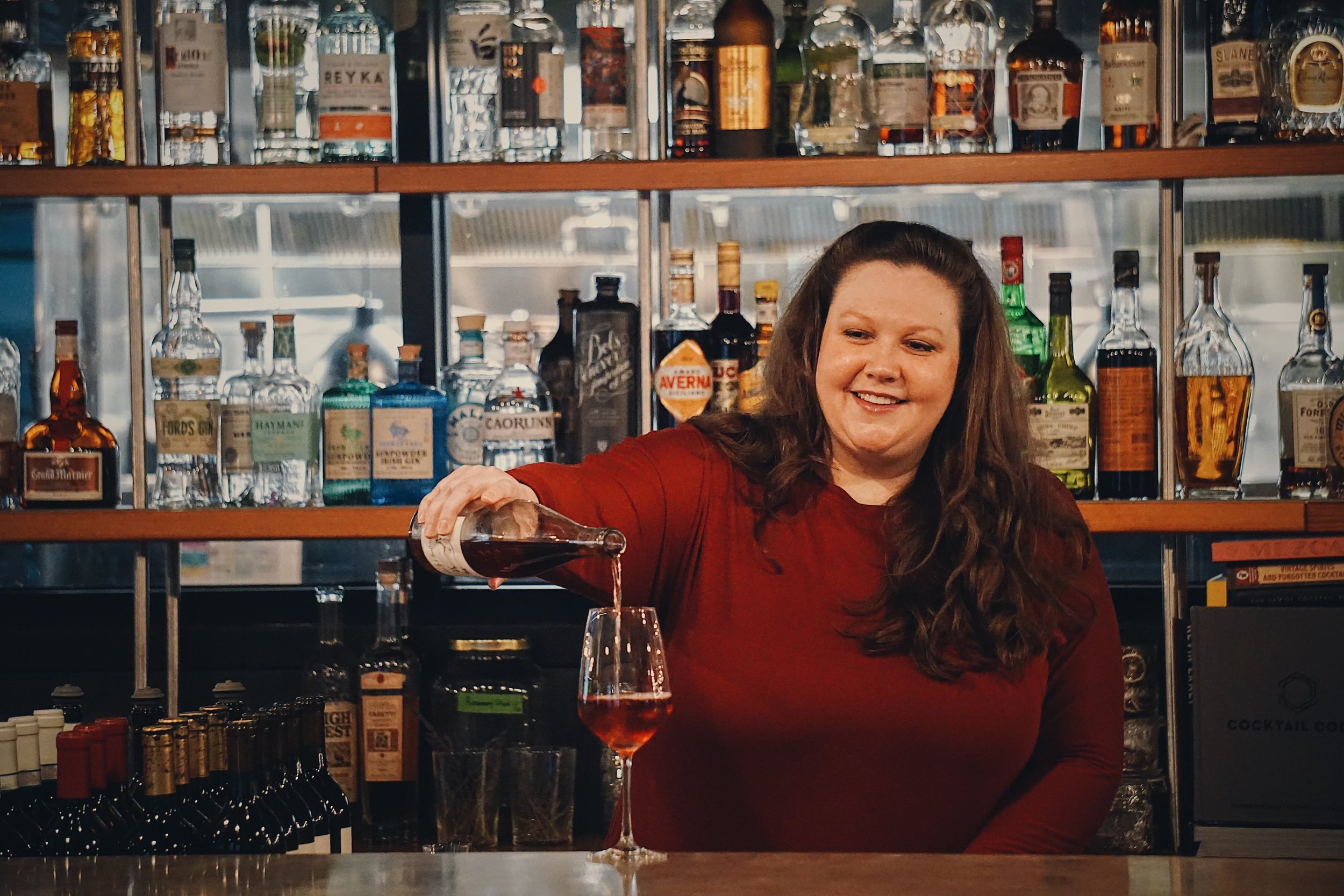 Amy stands behind a sleek, professional bar, expertly pouring a deep pink rosé into a wine glass