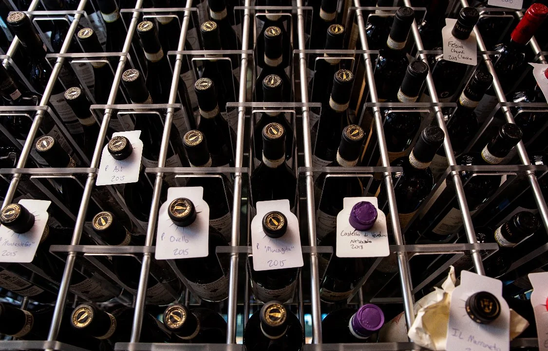 A dramatic, low-angle shot looking up at wine bottles resting on their sides in a professional cellar. Several bottles feature handwritten neck tags.