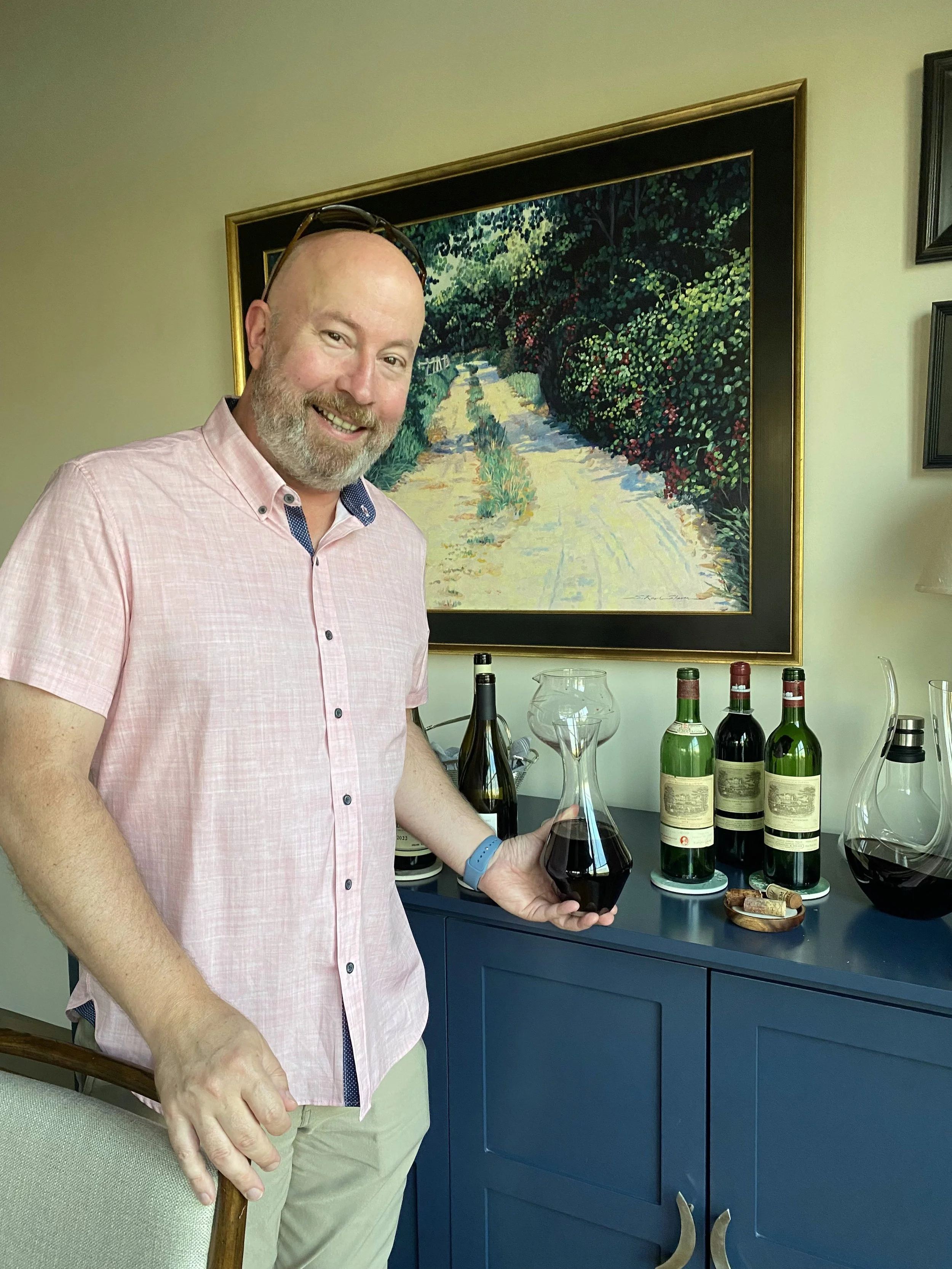 Wine enthusiast Kevin C. stands behind a row of open wine bottles, holding a striking red glass decanter.