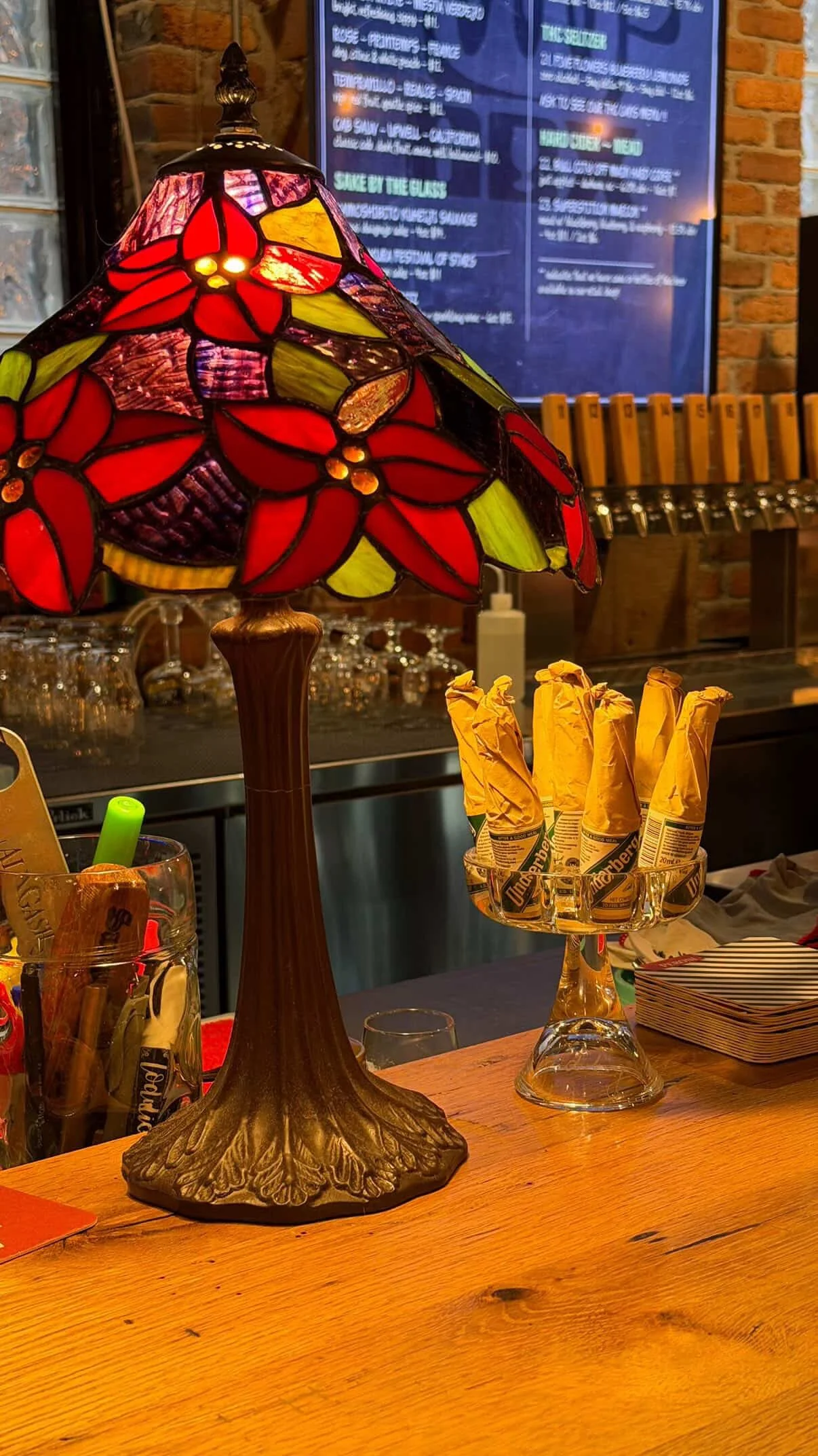 "A close-up detail shot of a decorative red stained-glass lamp illuminating a display of Underberg bitters. In the soft-focus background, a row of ten professional beer tap handles.