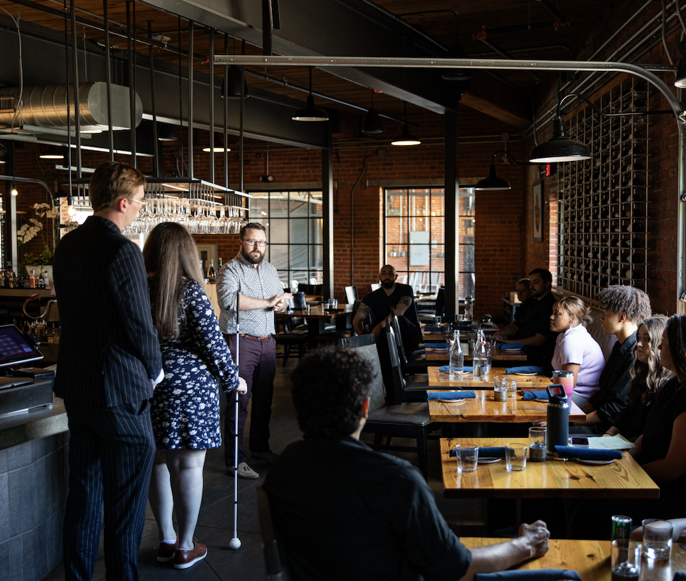 Certified Sommelier Amy stands confidently with her white cane while leading a wine education seminar. Accompanied by two assistant sommeliers, she speaks to a group of restaurant professionals about wine program standards and service.