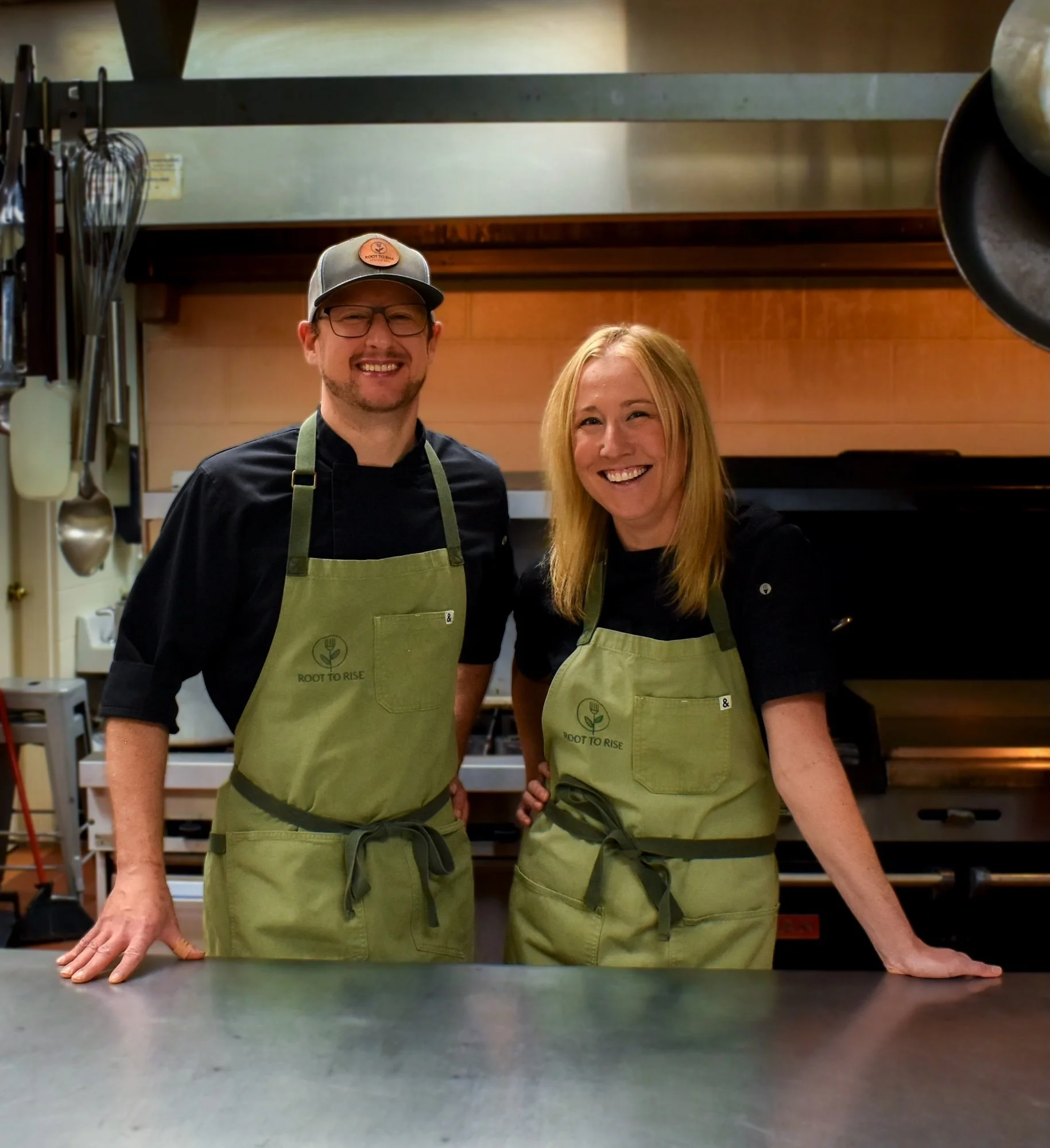 Chefs Jenn Cimino and Ryan Dunn of Root to Rise standing together in a professional commercial kitchen. They are wearing their signature green branded aprons