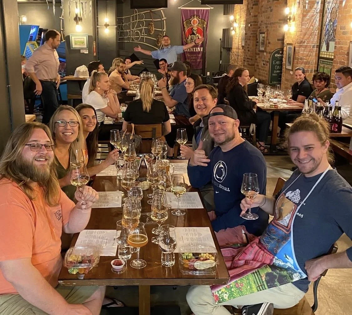 A vibrant group photo of 18 guests seated at a long table for a private wine tasting at GBX. Capturing the communal and joyful spirit of Amy’s events