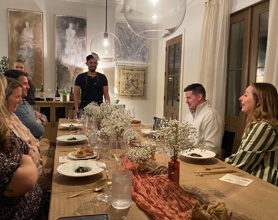 A private chef stands at an elegantly set dining table, sharing the story of the meal with a family in their home. The table is beautifully decorated, creating a warm and sophisticated atmosphere for an intimate, plated dining experience.