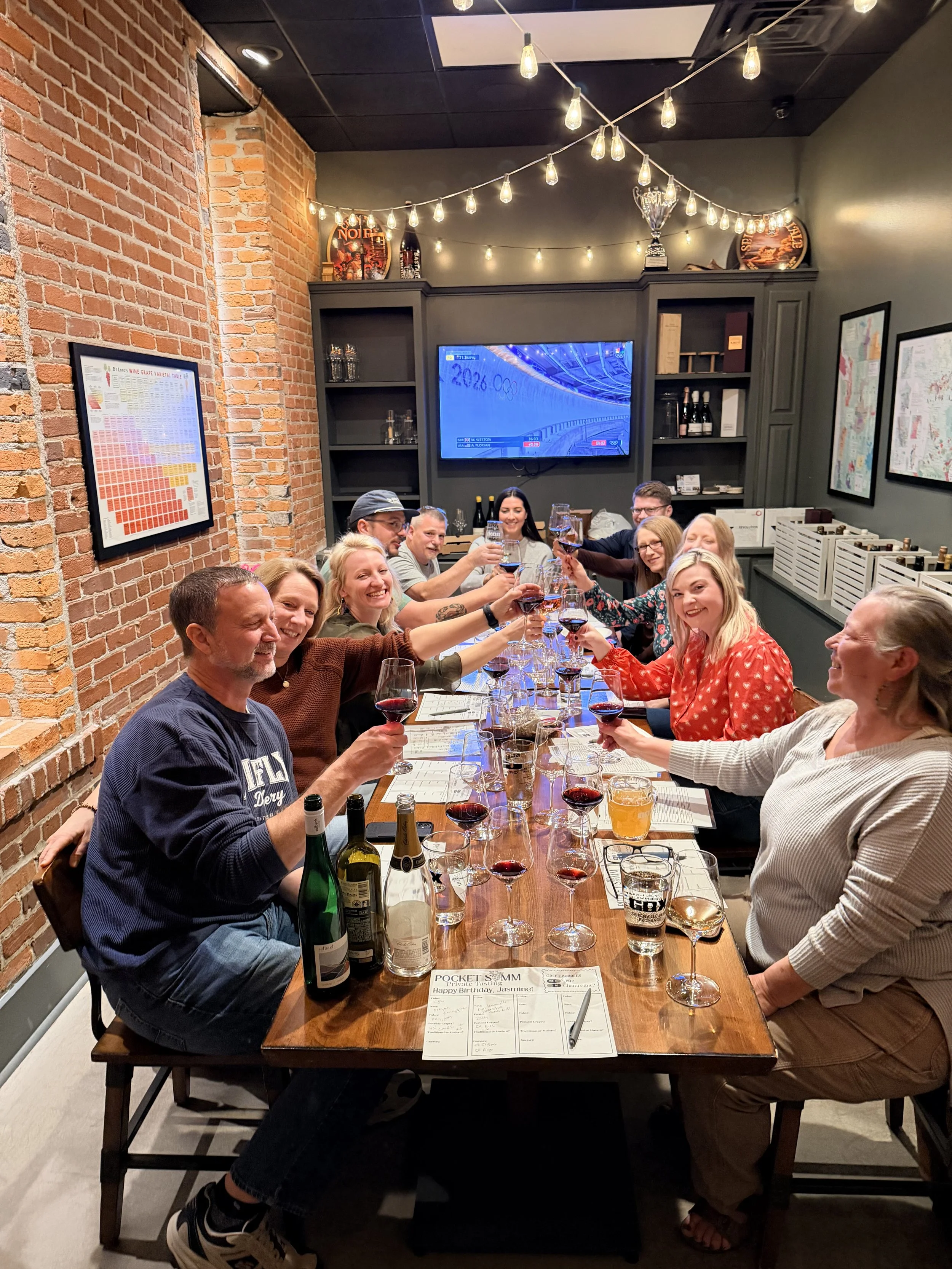 A lively group of twelve guests raising their glasses for a toast during a private wine tasting in the GBX boardroom. The table is filled with wine flights and pizzas.