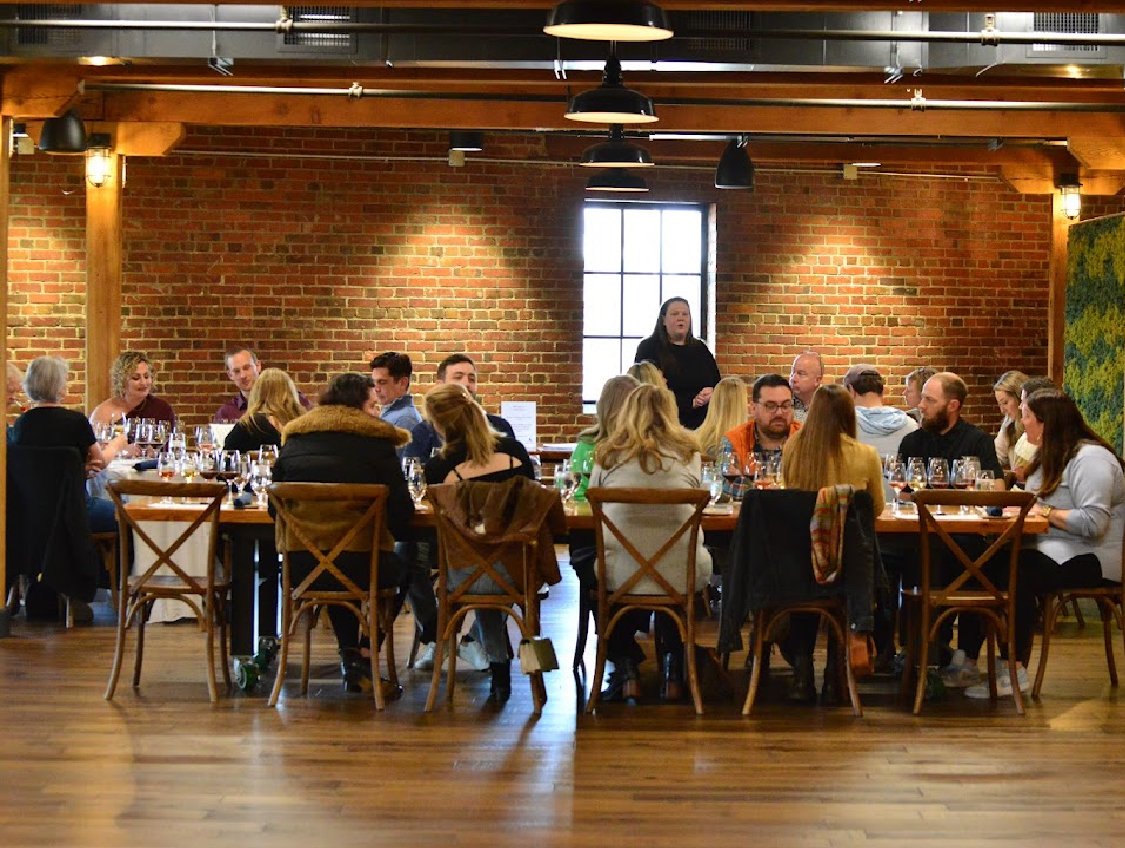 Certified Sommelier Amy stands at the front of a room, expertly leading a private tasting for 30 guests. Each person has a flight of four organic wines and a curated charcuterie board, enjoying an educational and social wine experience.