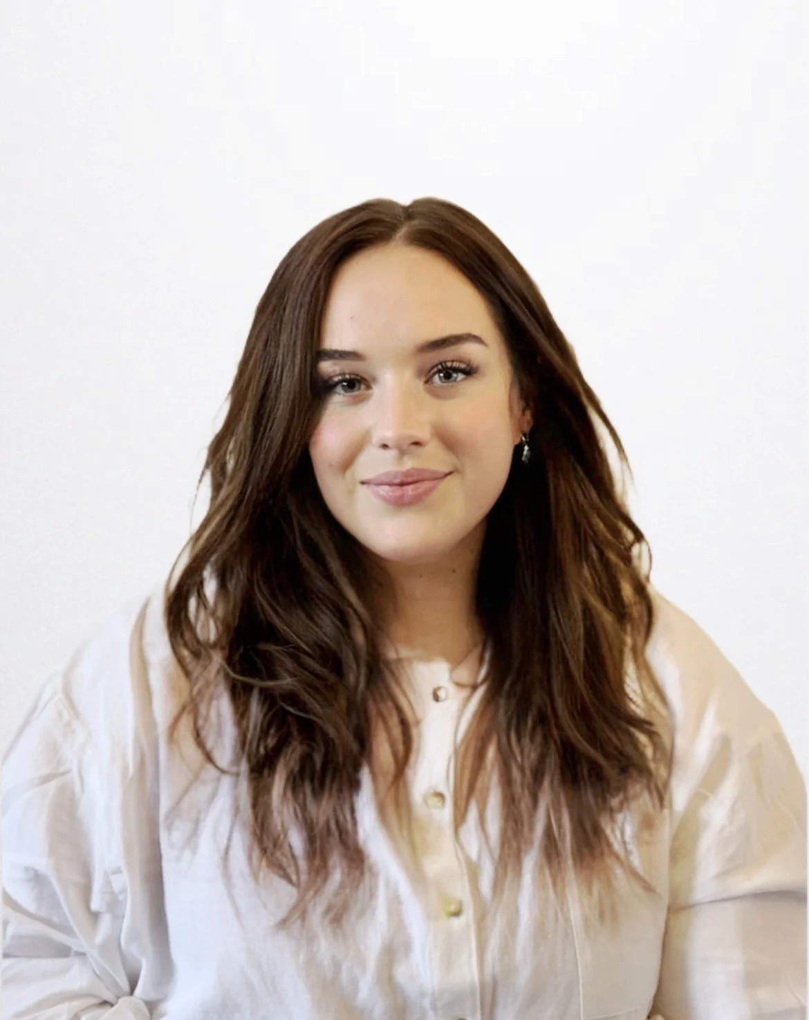 A young woman with long, wavy brown hair, light skin, and wearing a white blouse, posing against a plain white background.
