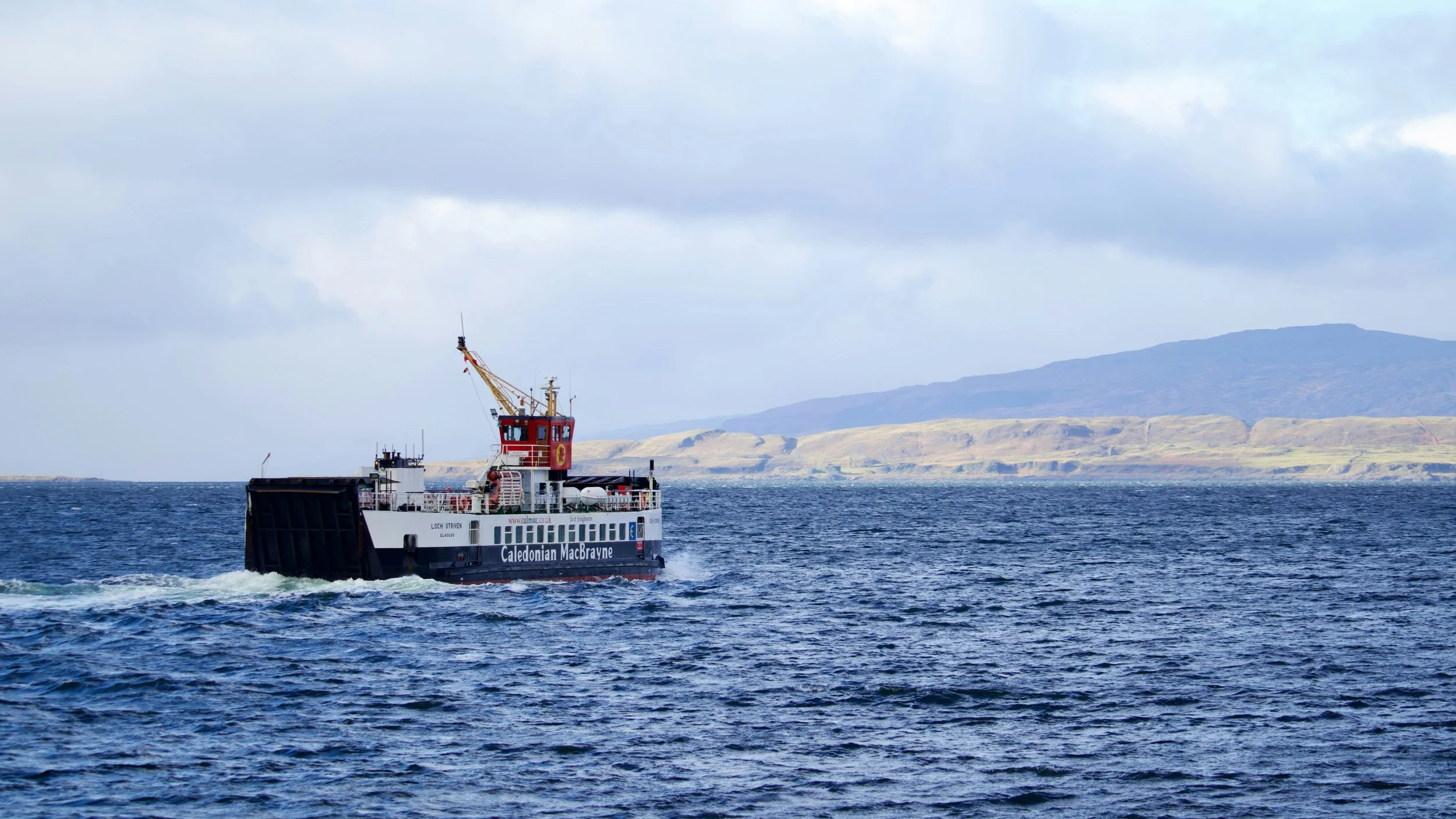 A ferry boat named Caledonian MacBrayne sailing on the ocean with hills in the background and cloudy sky above.