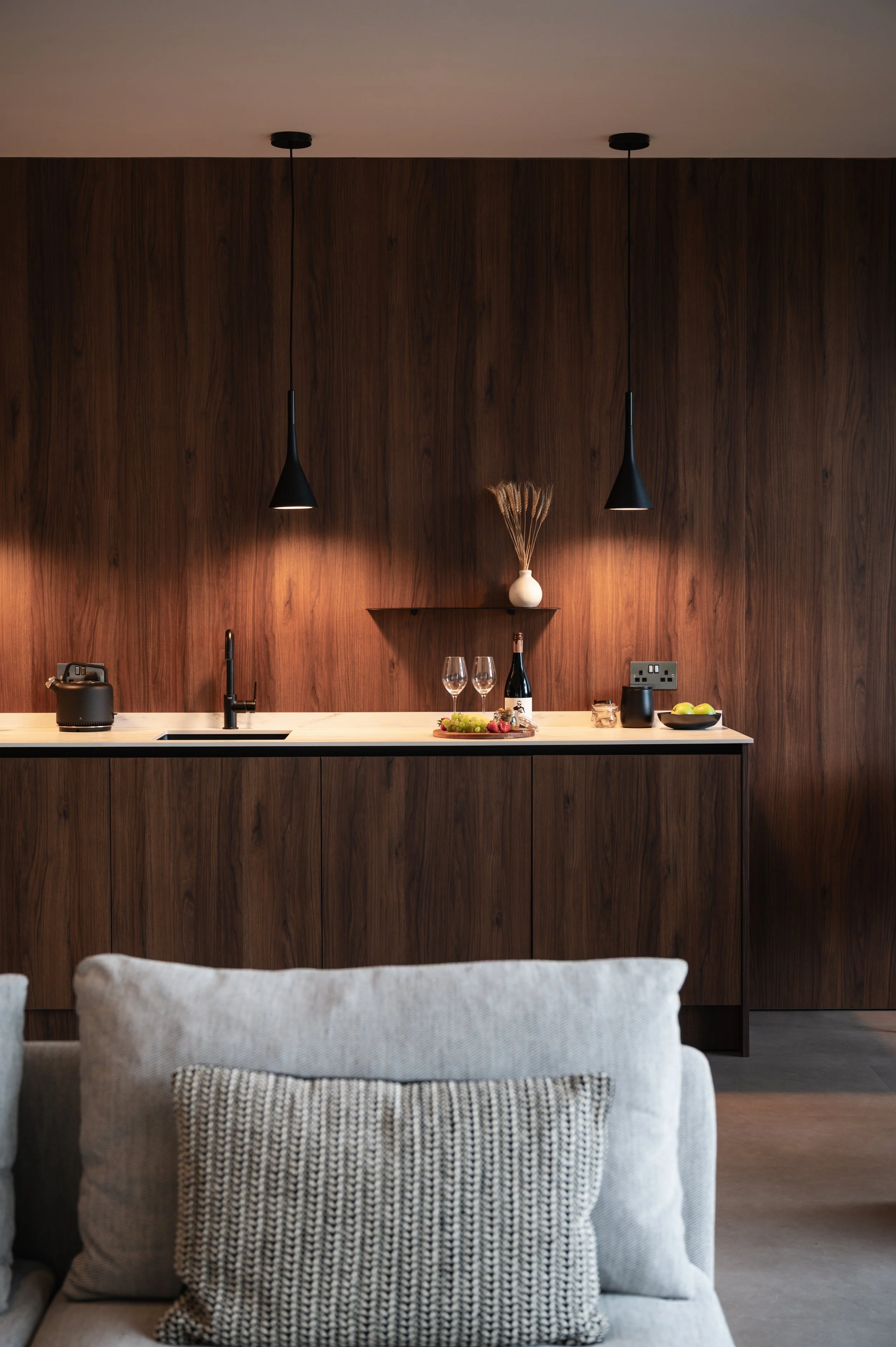 Modern kitchen with dark wood cabinets, black pendant lights, and a white countertop. Decor includes a white vase with dried flowers, wine glasses, a bottle of wine, a bowl of fruit, and small appliances. A beige armchair with a textured pillow is in the foreground.