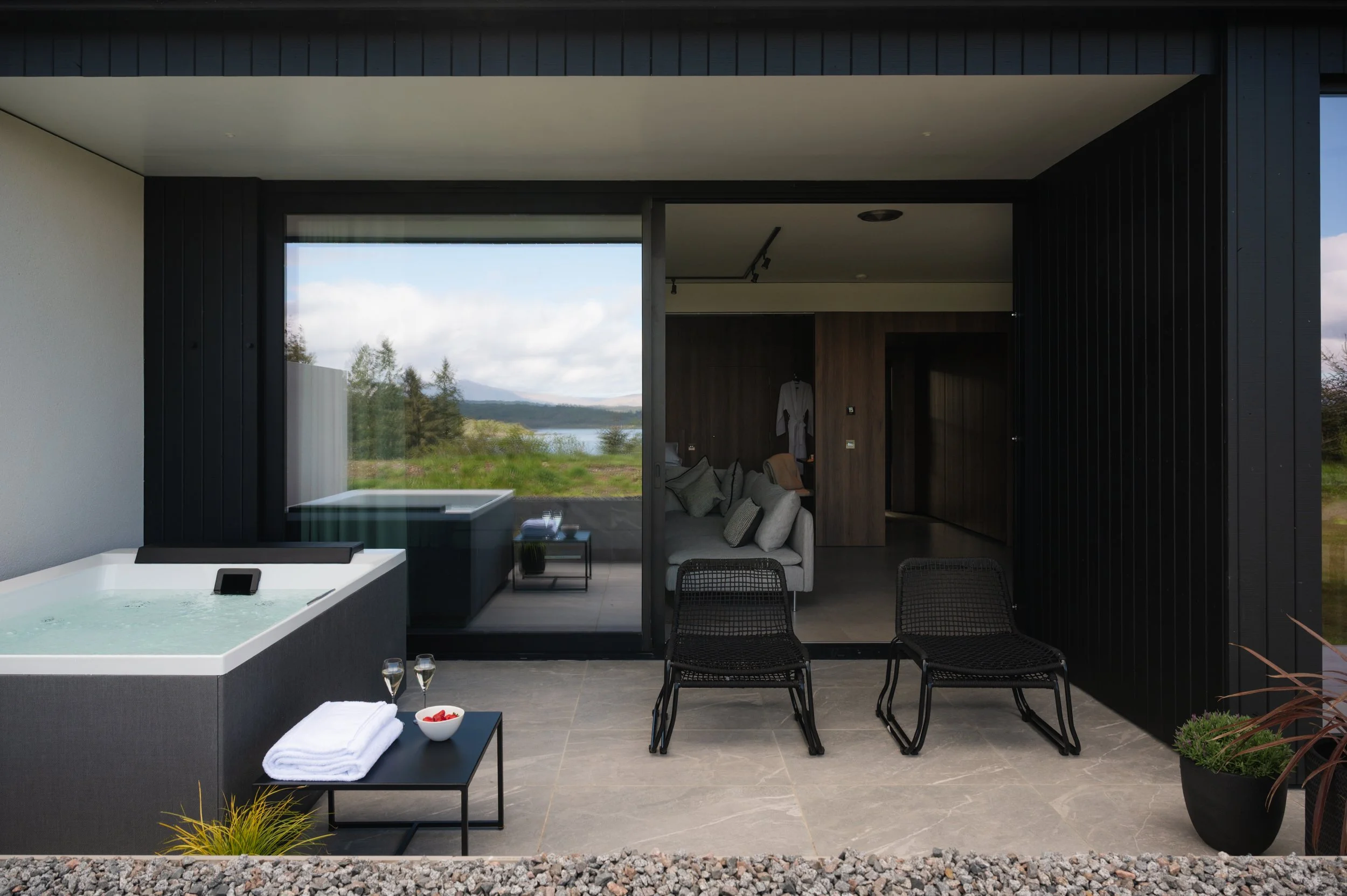 Modern outdoor patio area with a hot tub, black chairs, a small table with towels and snacks, and a view of nature through large glass doors, leading to a living room with a sofa and wood-paneled wall.