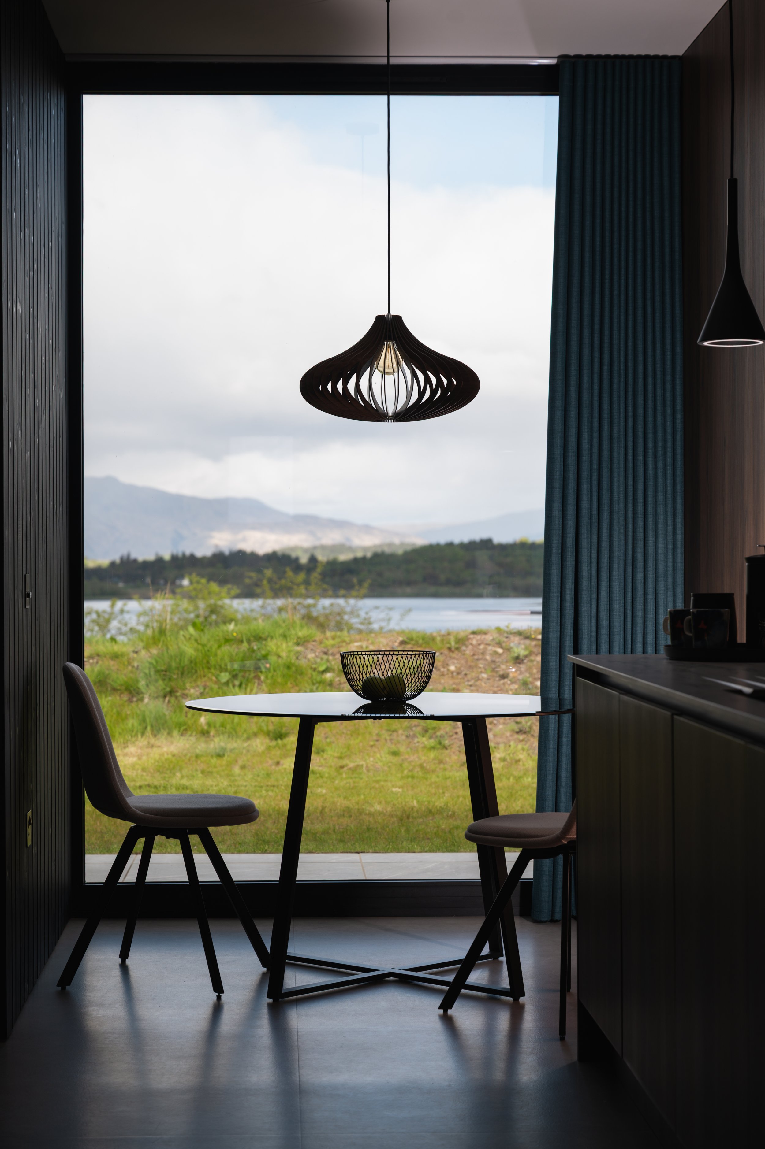 Interior view of a dining area with two chairs, a round table with a decorative bowl, and large glass sliding doors opening to a landscape with body of water, grassy area, trees, and hills under a cloudy sky. A hanging lamp is centered above the table, and heavy blue curtains frame the window on the right.