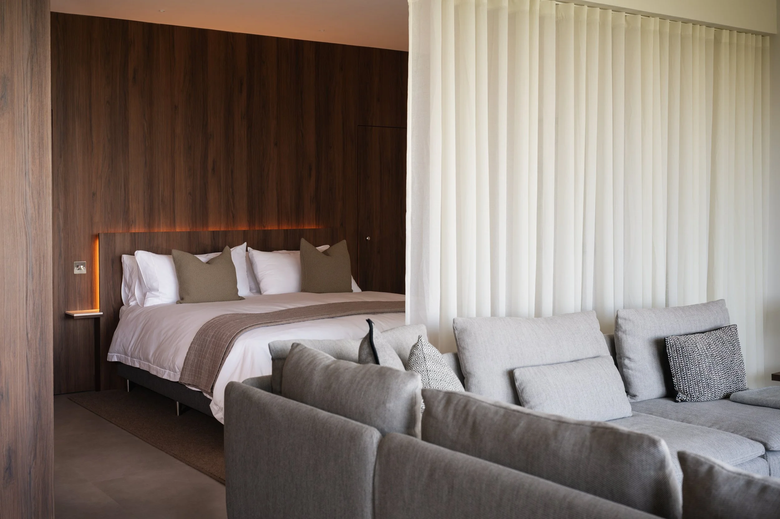 A bedroom with a large bed featuring white sheets, beige and gray pillows, and a beige blanket. The headboard wall is dark wood with warm lighting, and there is a white curtain on the right side, along with a seating area with a gray armchair and patterned pillows.
