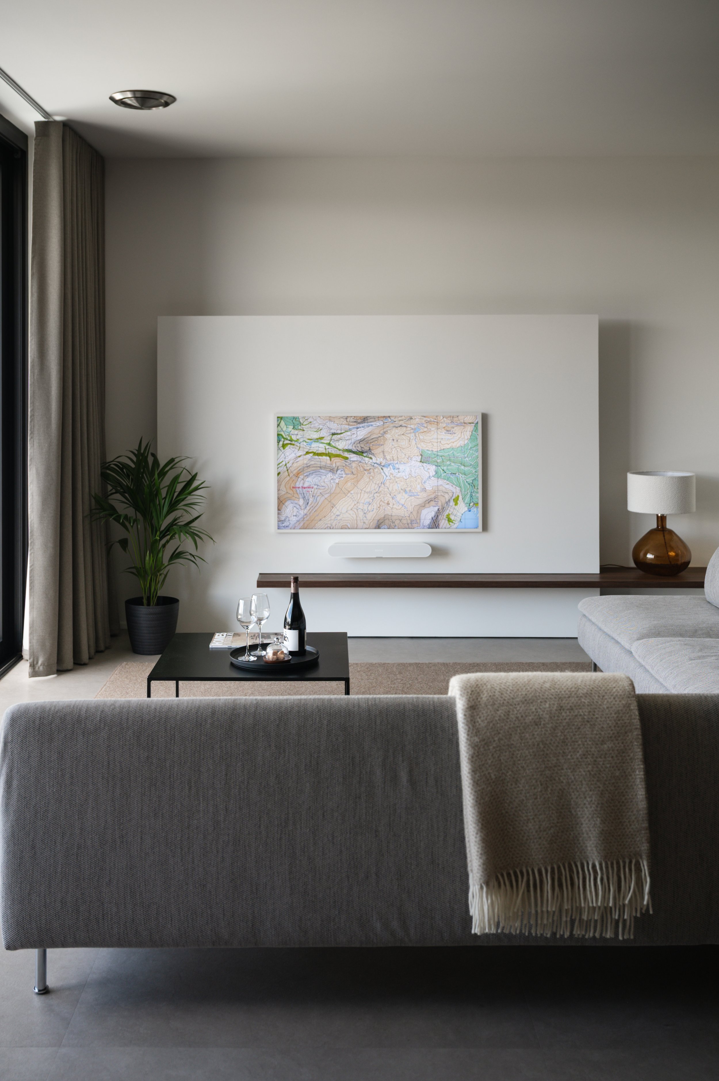 Living room with beige sofa, black coffee table with a bottle of wine and glasses, potted plant, wall-mounted TV displaying a map, side table with lamp, curtains, and neutral decor.