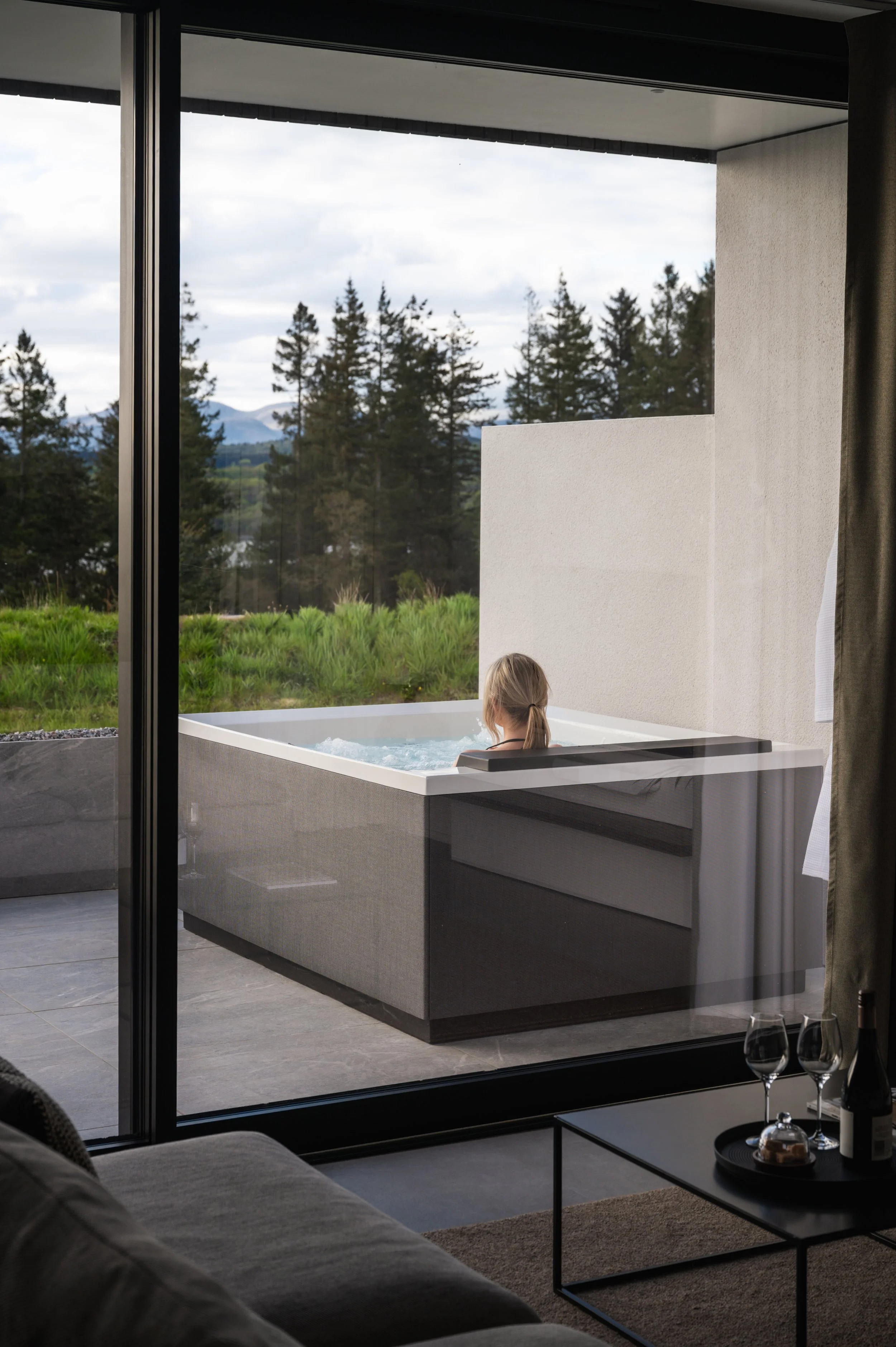 Person relaxing in a jacuzzi on a balcony with a view of trees and mountains