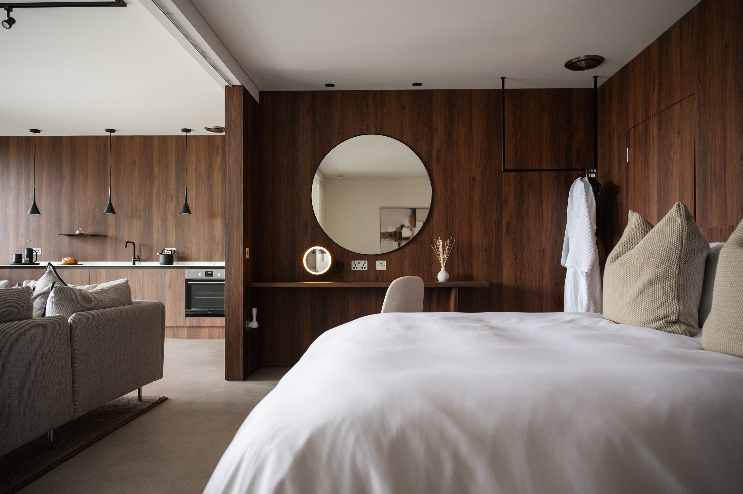 Interior of a modern bedroom with a wood-paneled wall, a bed with beige pillows, a round mirror, and a small vanity area, adjacent to an open-plan kitchen with pendant lights.