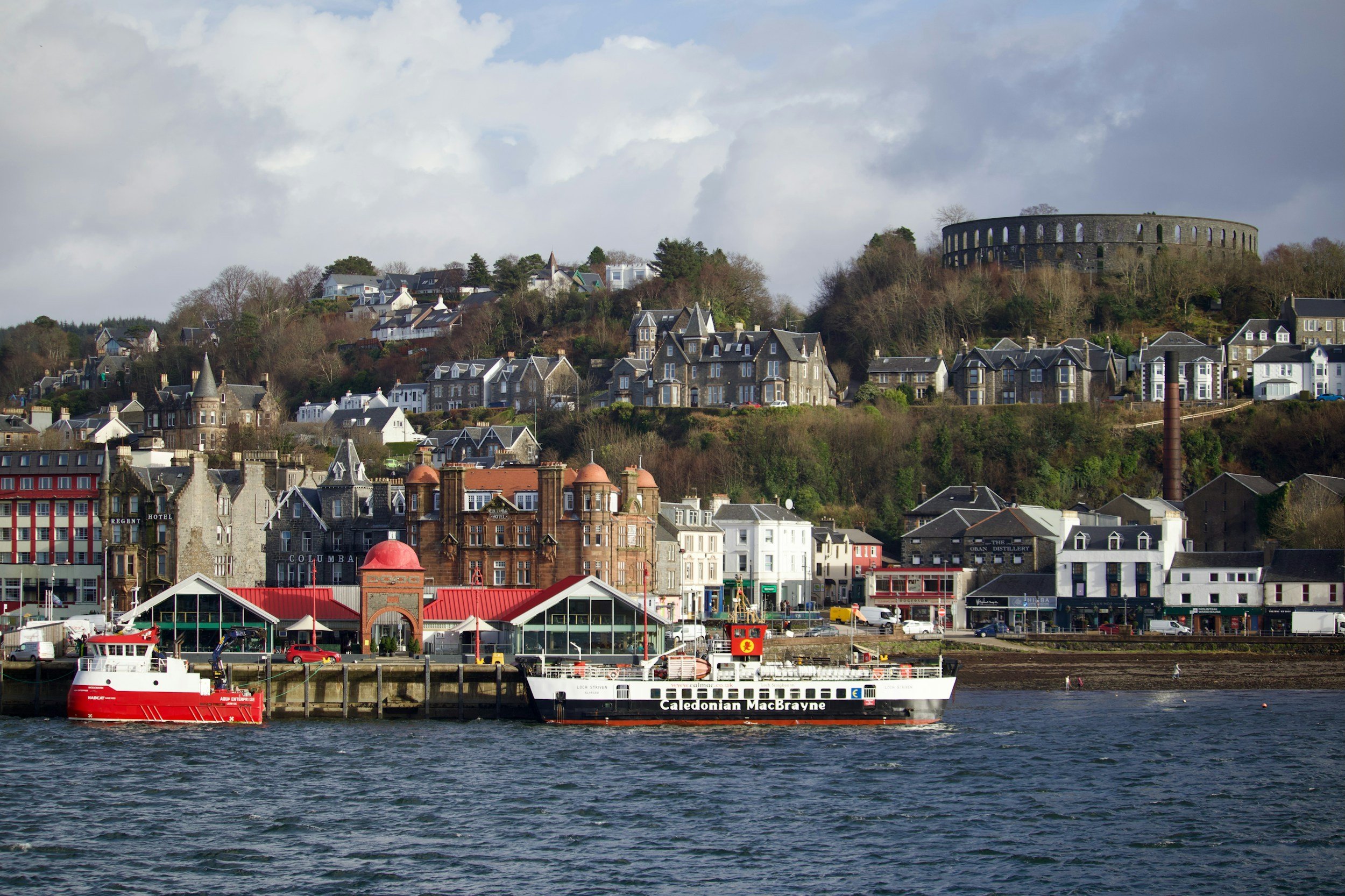 A harbor scene with two ferries on the water, there are buildings along the shoreline and houses on a hill in the background, with a large circular structure on the hilltop.